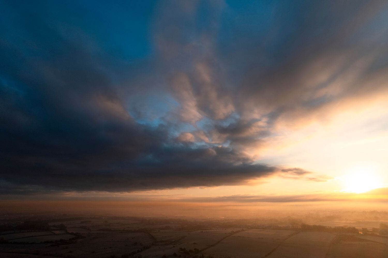 Dramatic sunset sky with dark storm clouds over a misty rural landscape at dusk.