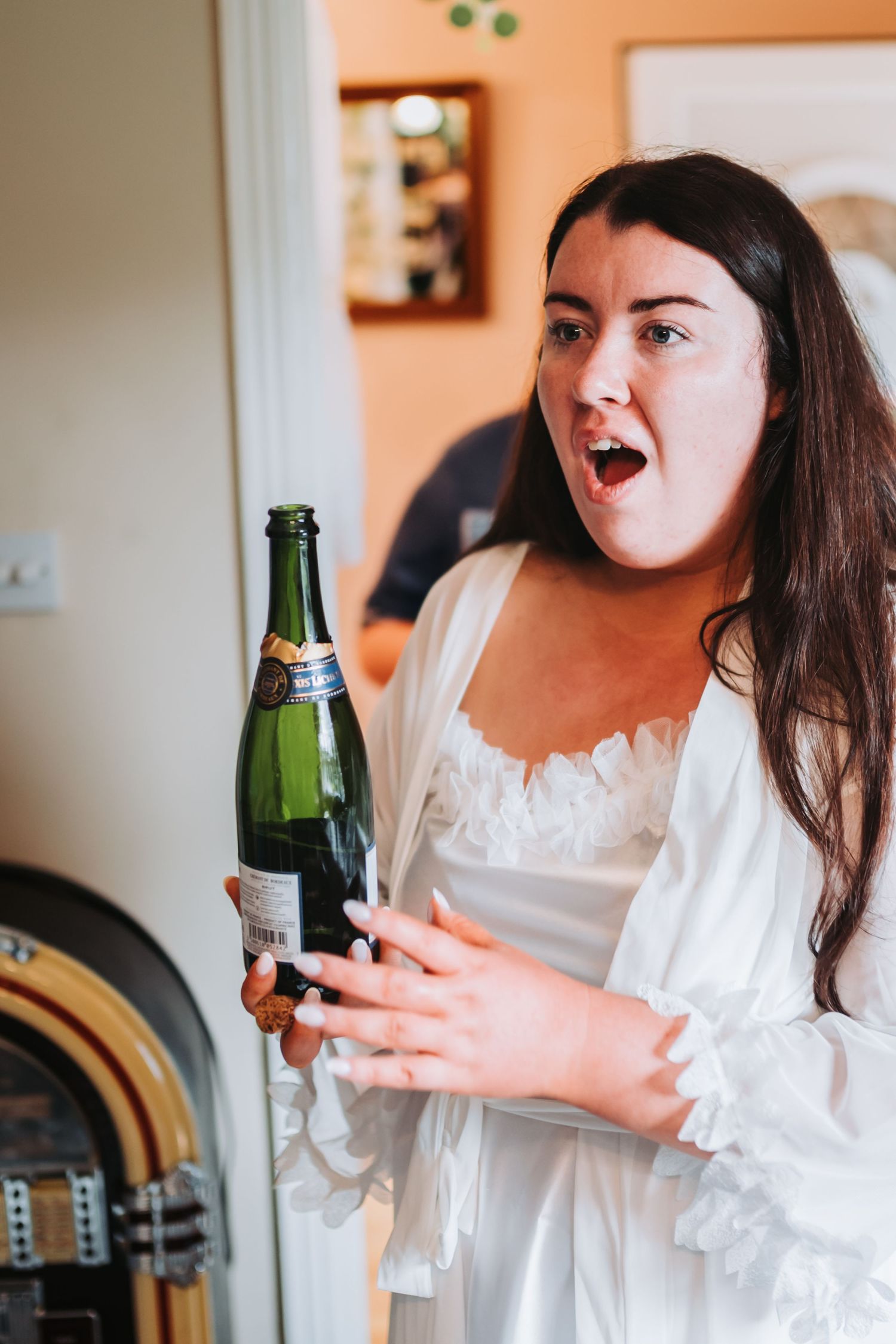 Excited bride in white robe holding champagne bottle during wedding morning preparations.