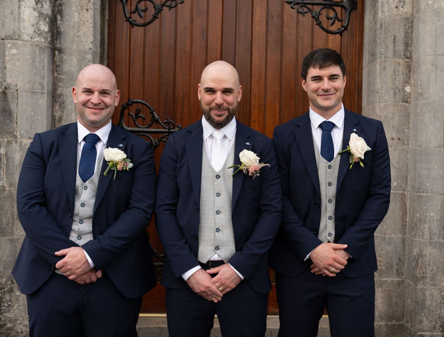 Three groomsmen in matching navy suits with grey waistcoats and white rose boutonnieres pose at church doors.