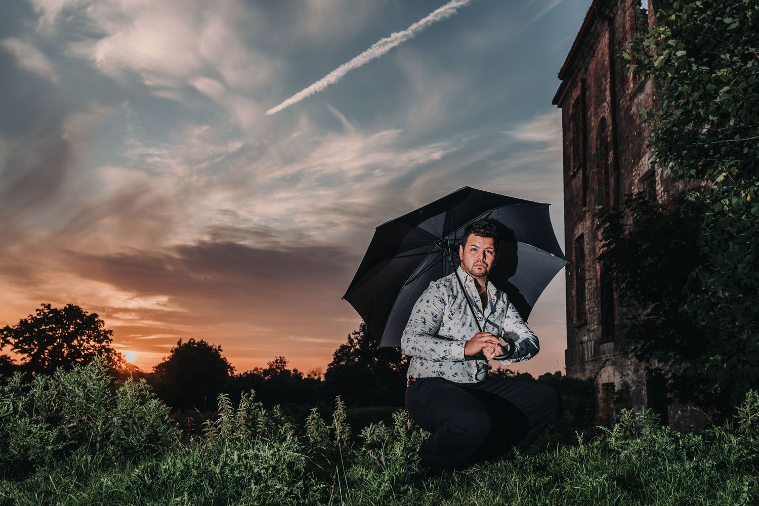 Man crouching with black umbrella at sunset near old building, holding camera, dramatic cloudy sky backdrop.