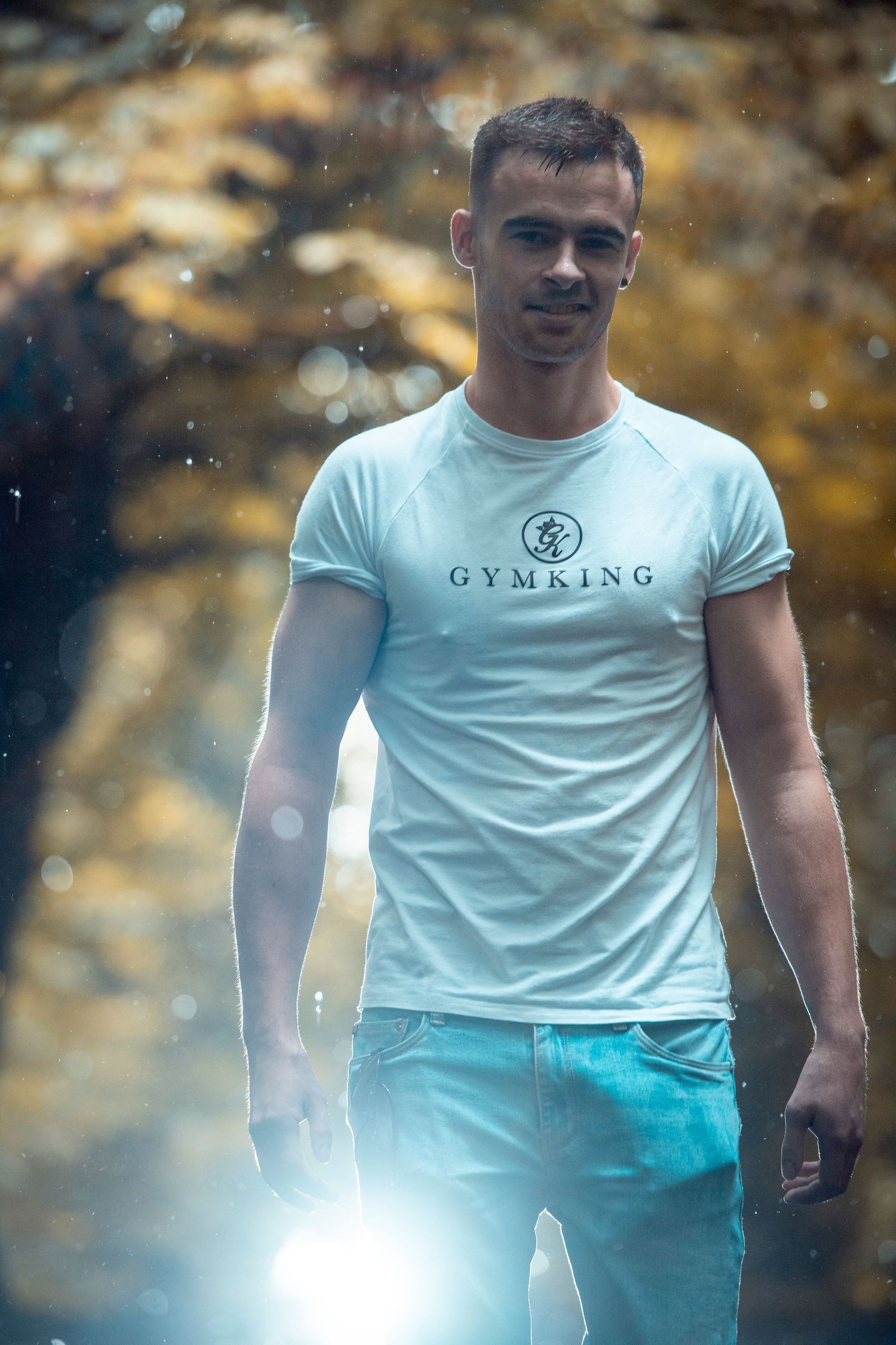 Muscular man wearing a white Gym King t-shirt and jeans standing in an autumn forest with bokeh lighting effects.