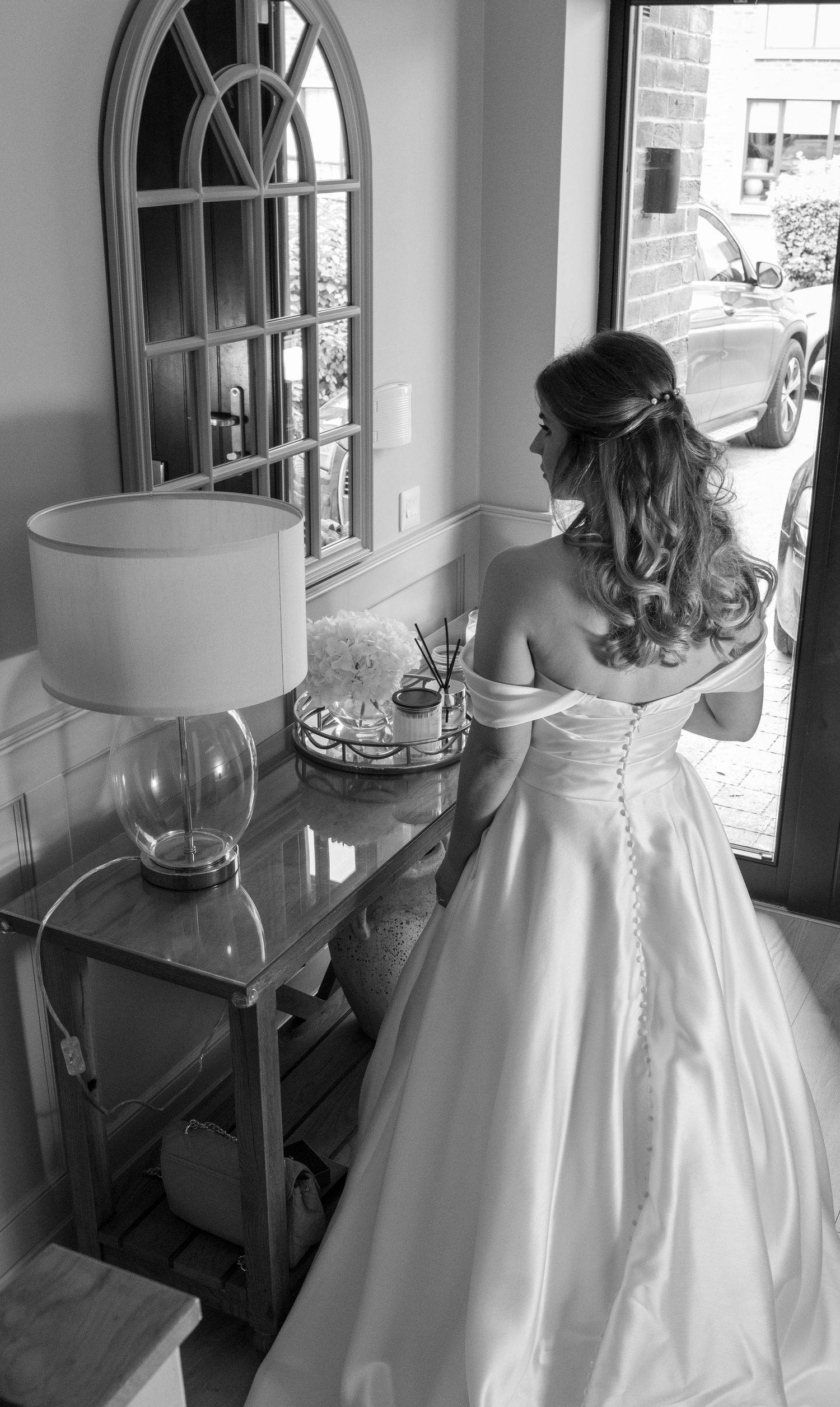 Bride in off-shoulder white wedding gown viewed from behind, standing near a console table with lamp and arched mirror.