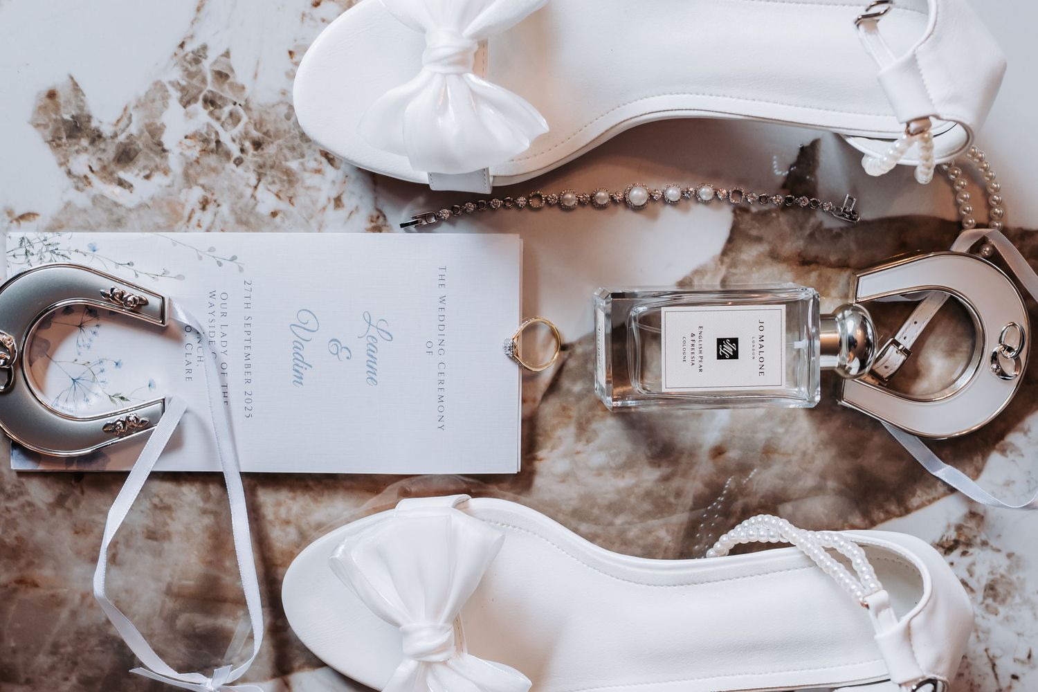 Elegant wedding flat lay featuring white bridal heels, Jo Malone perfume, pearl jewelry, and invitation on marble surface.