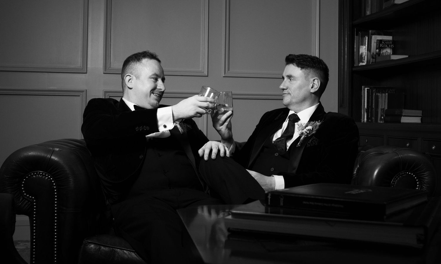 Two men in tuxedos share a toast while seated on a leather sofa in a classic black and white portrait.
