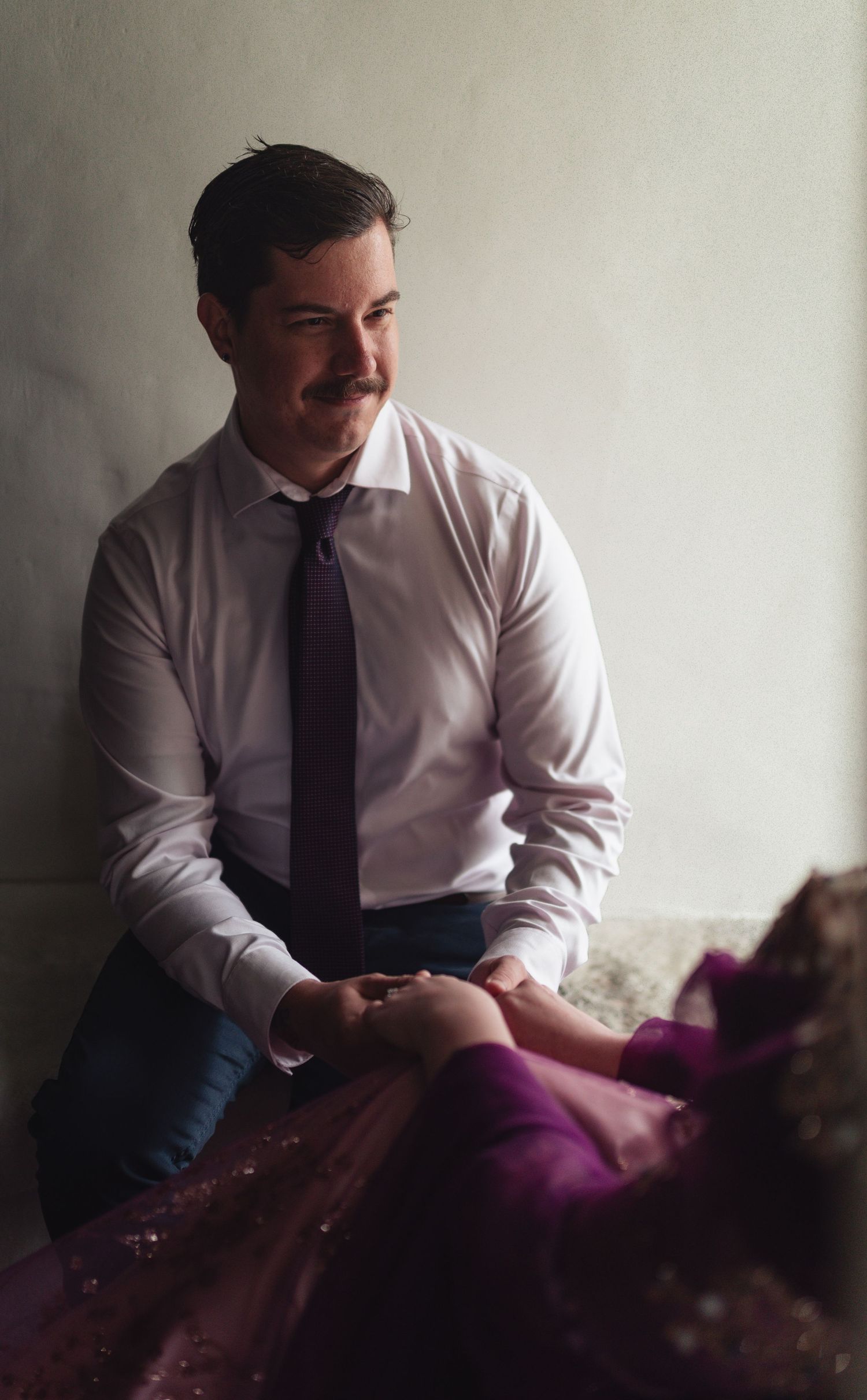Groom in white shirt and burgundy tie sits holding brides purple tulle gown during wedding day.