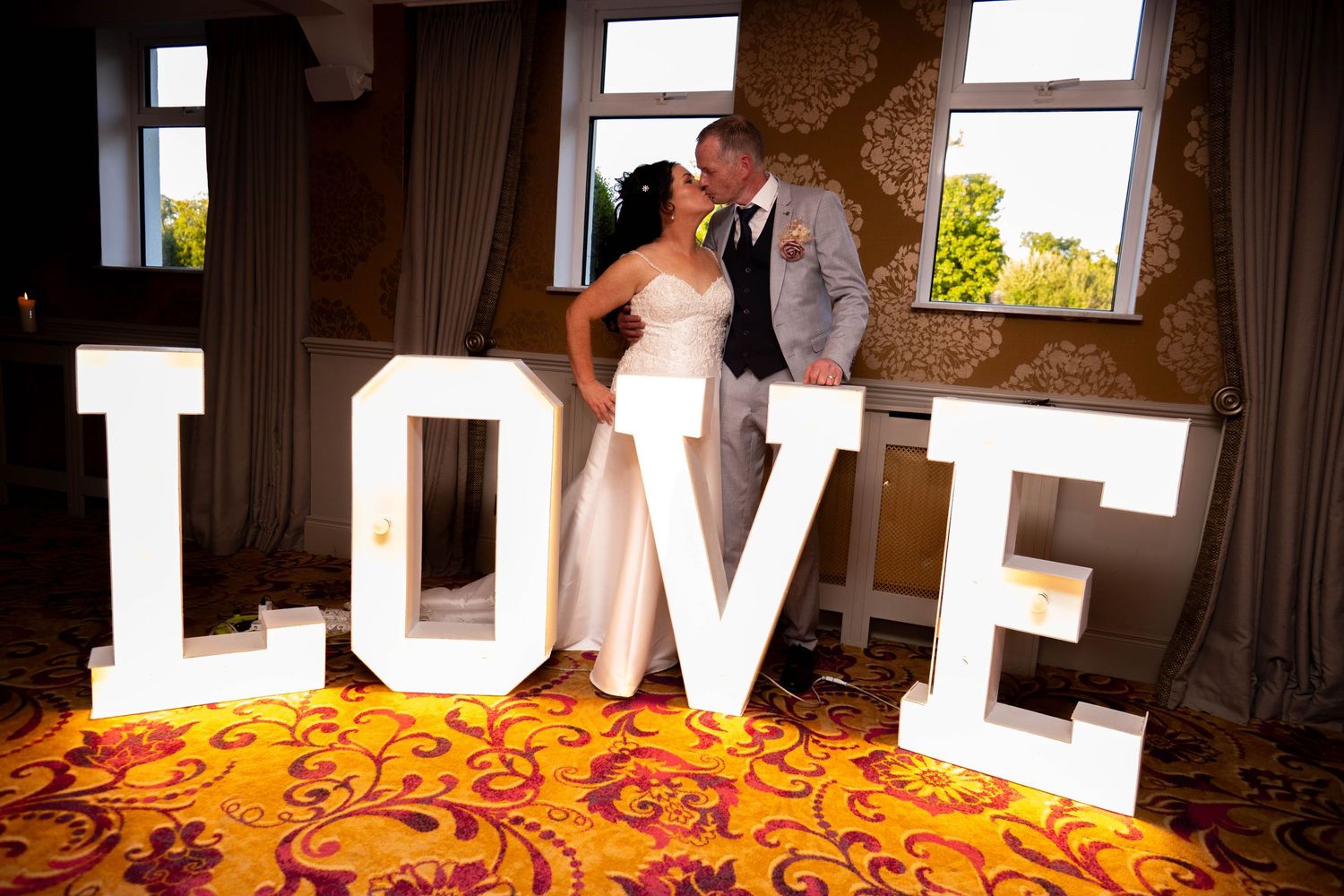 Bride and groom kissing behind illuminated LOVE letters on a decorative dance floor indoors.