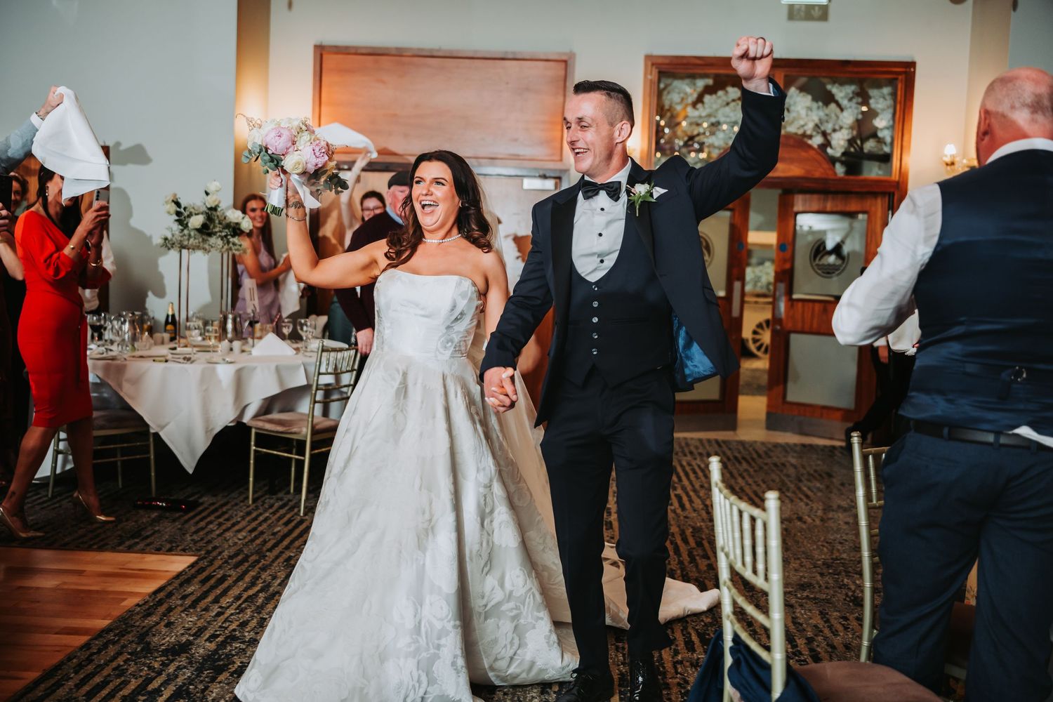 Bride and groom celebrate joyfully at wedding reception entrance, bride in white gown, groom in black tuxedo.
