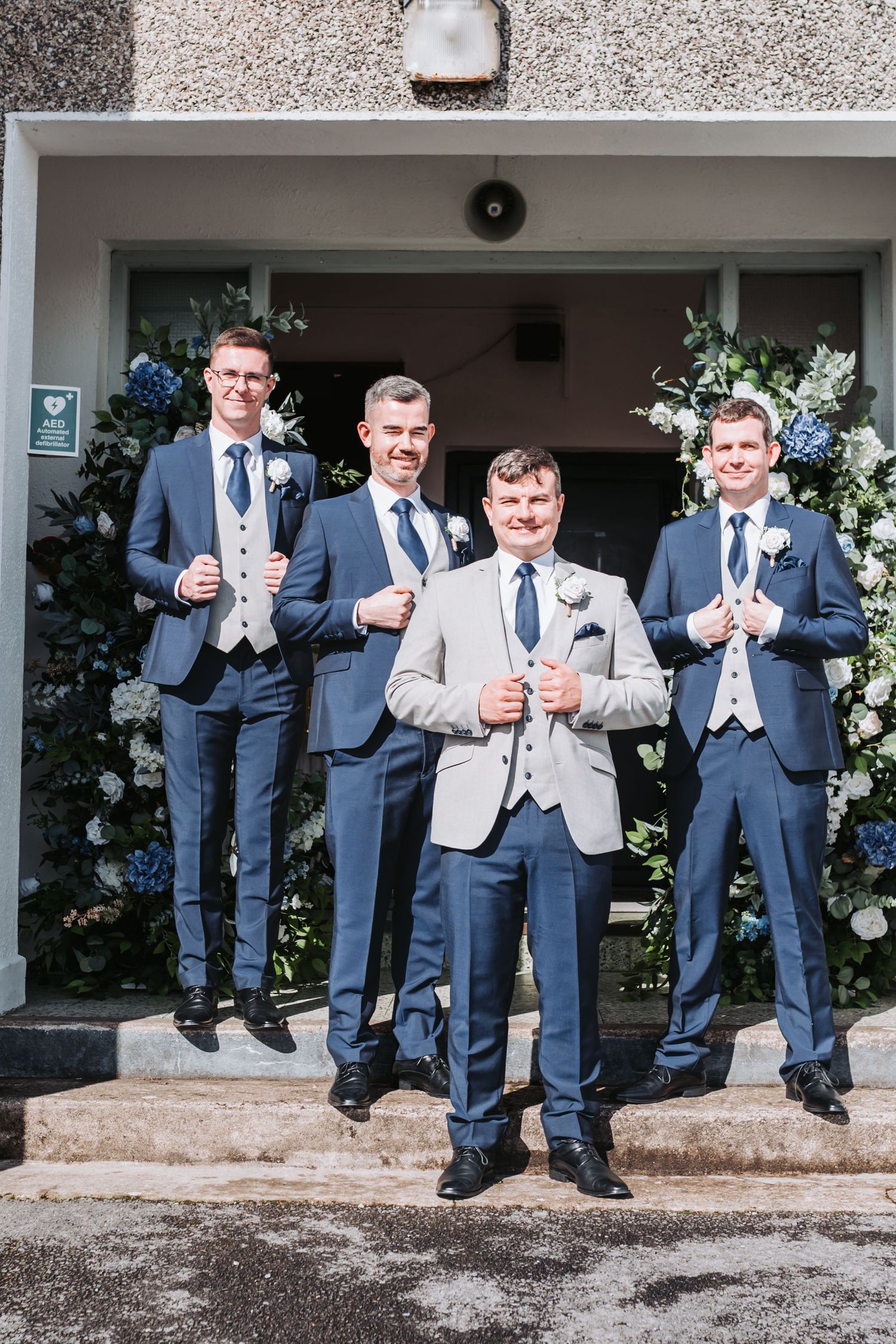 Groom in beige suit poses with three groomsmen in navy suits outside flower-decorated church entrance.