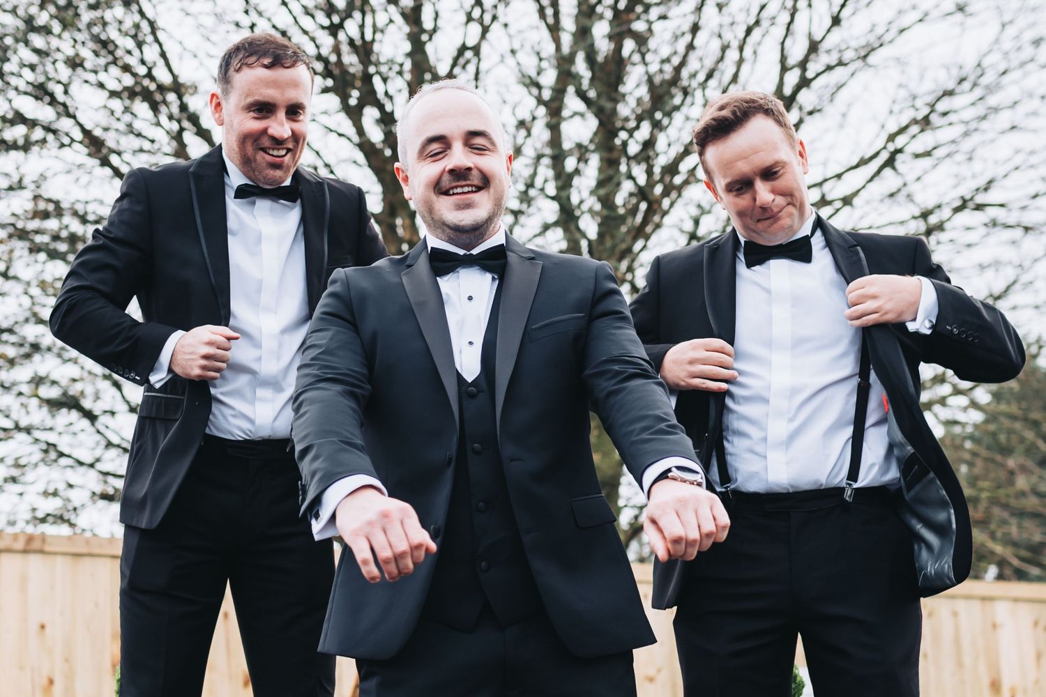 Three groomsmen in black tuxedos and bow ties posing outdoors, showing off their cufflinks with big smiles.