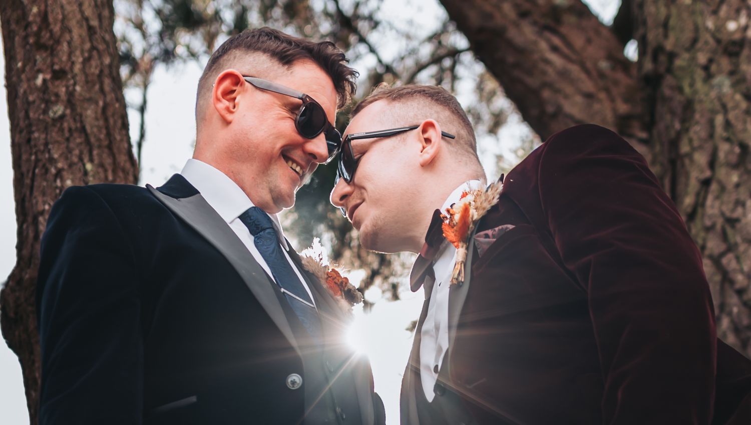 Two grooms in sunglasses share an intimate moment outdoors on their wedding day, with sunlight glowing between them.