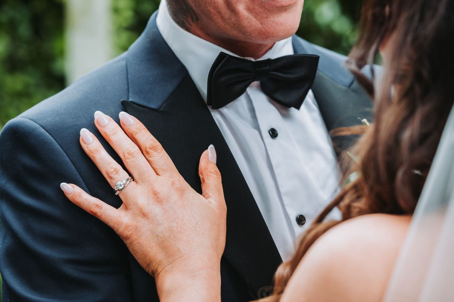 Close-up of bride's hand with diamond engagement ring resting on groom's black tuxedo lapel at wedding.