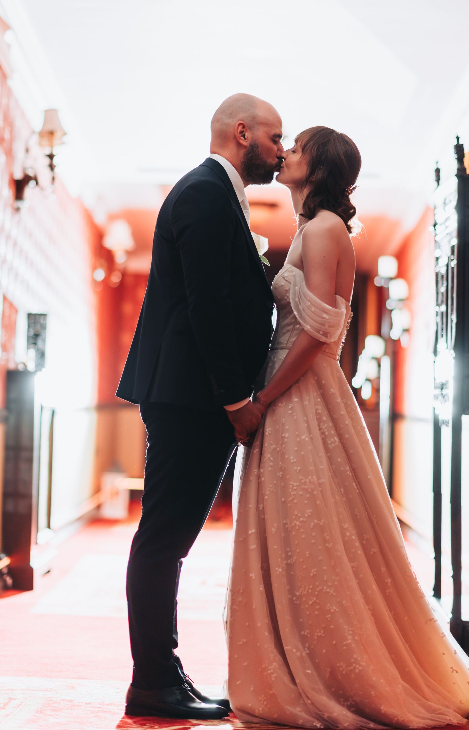 Bride and groom share a romantic kiss in a grand hallway, bride wearing a blush off-shoulder ballgown.
