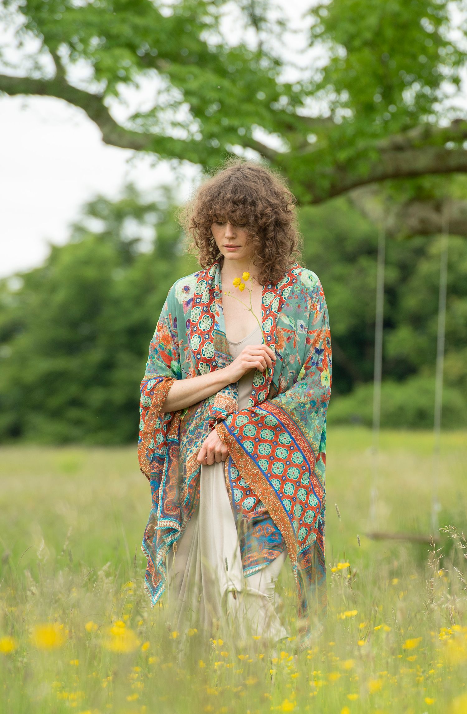 Woman in colorful boho kimono holding yellow flower in green meadow.