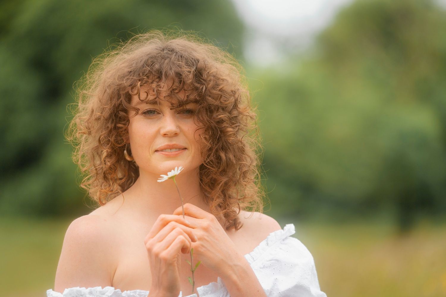 Curly-haired woman in white off-shoulder dress holding a daisy flower outdoors in a green field.