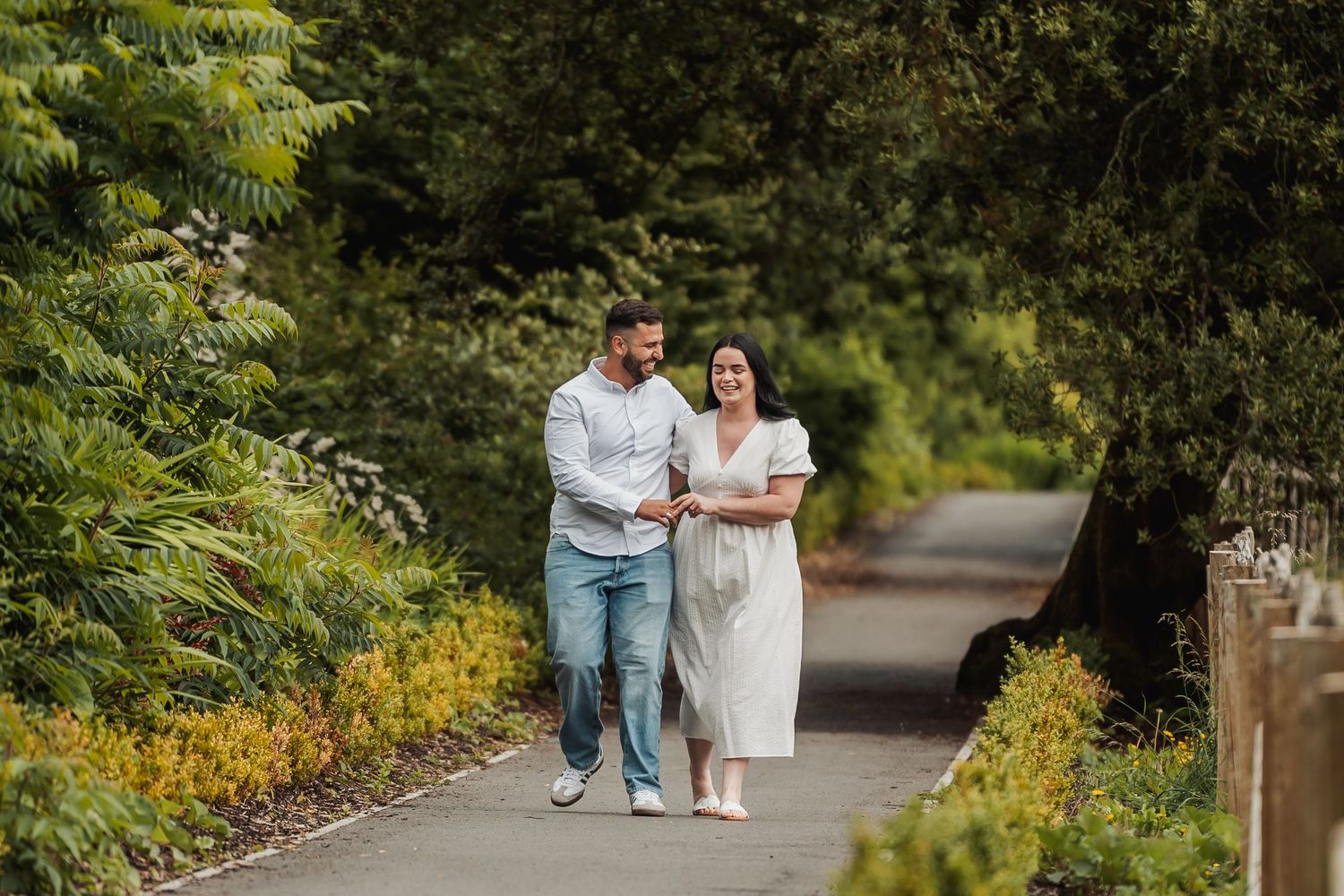 Couple walking hand-in-hand on a lush green garden path, smiling together on a sunny day.