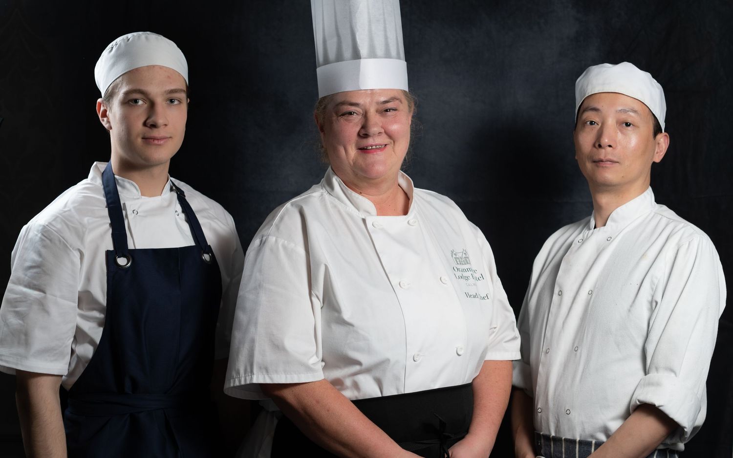 Three professional chefs in white uniforms and hats posing together against a dark background.