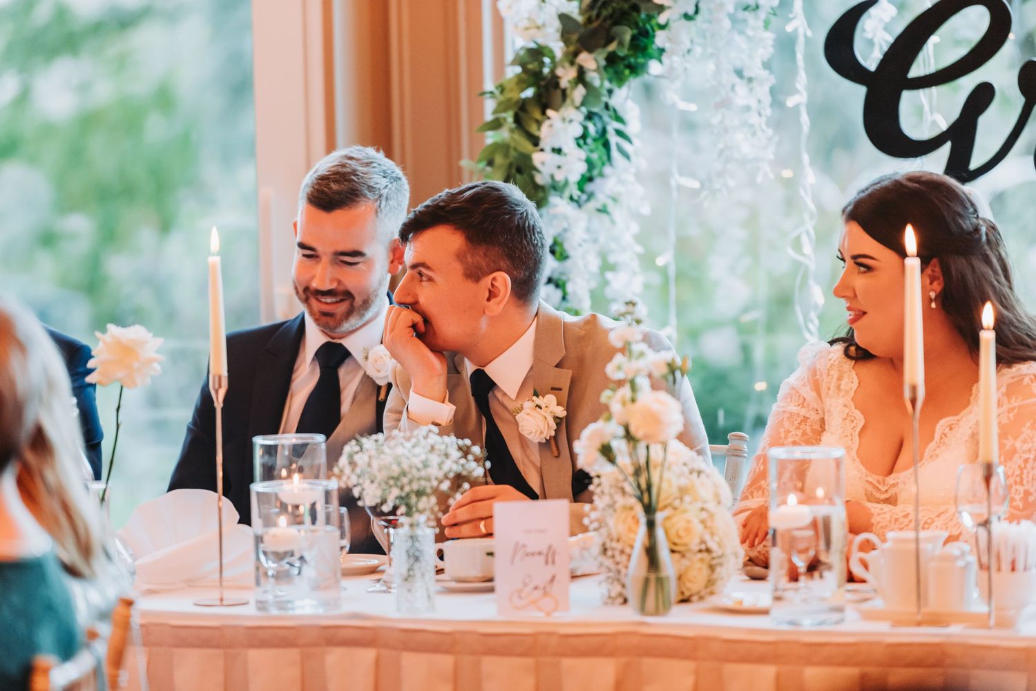 Groom and groomsman laughing together at elegant candlelit wedding reception head table.