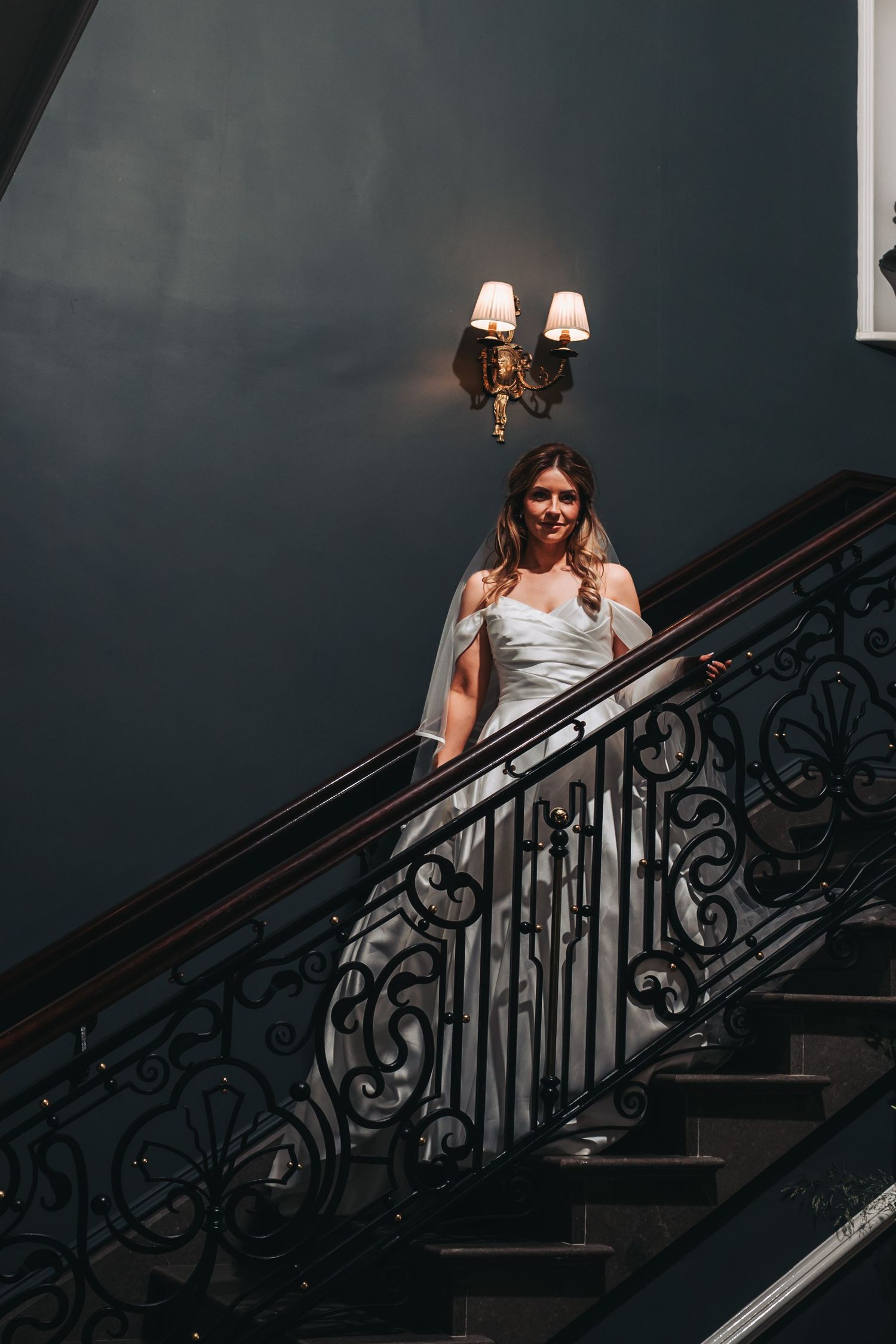 Elegant bride in white gown descending ornate iron staircase under warm wall sconce lighting.