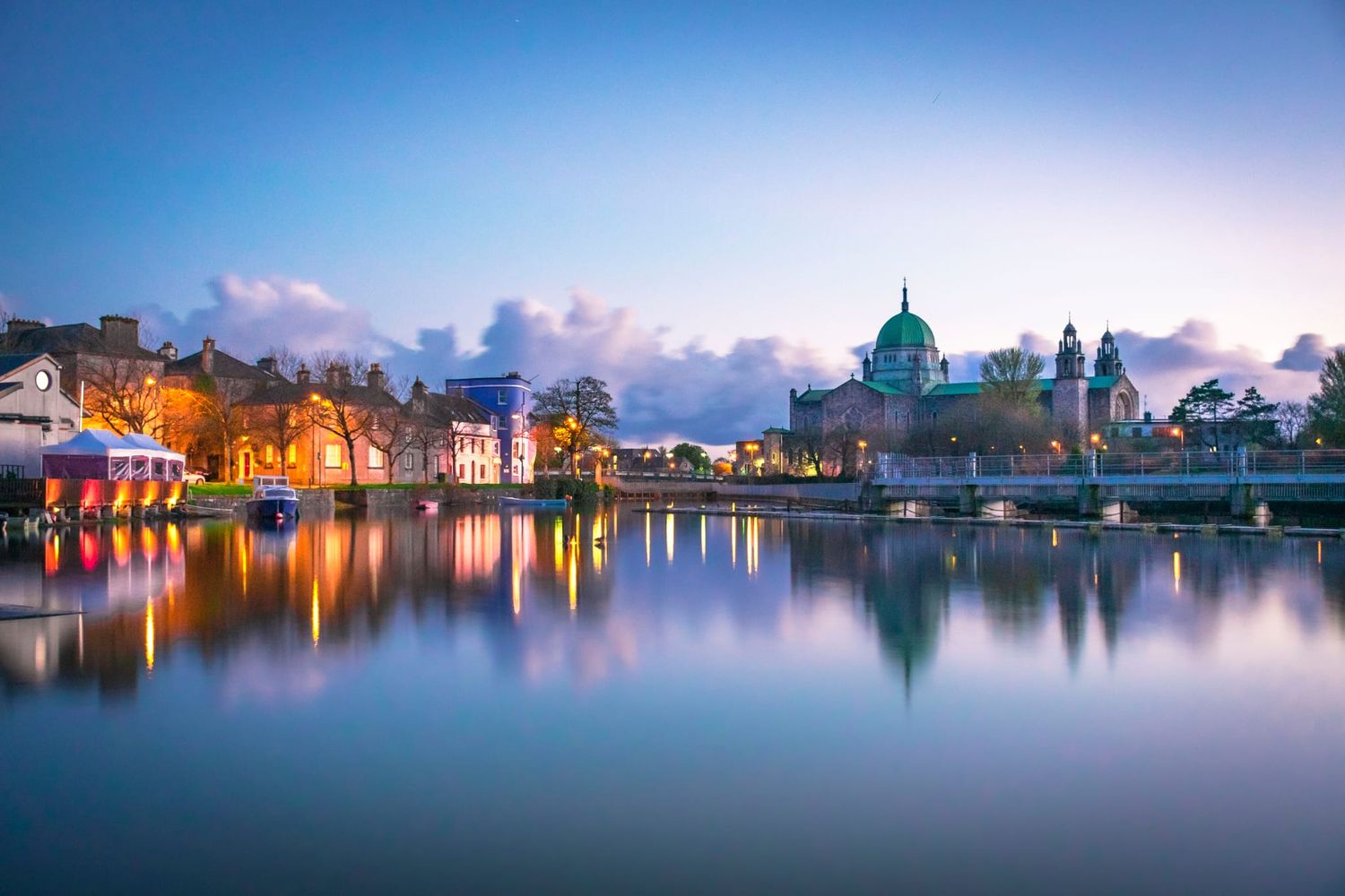 Panoramic view of illuminated buildings reflecting in calm waters during twilight with vibrant blue and orange hues.