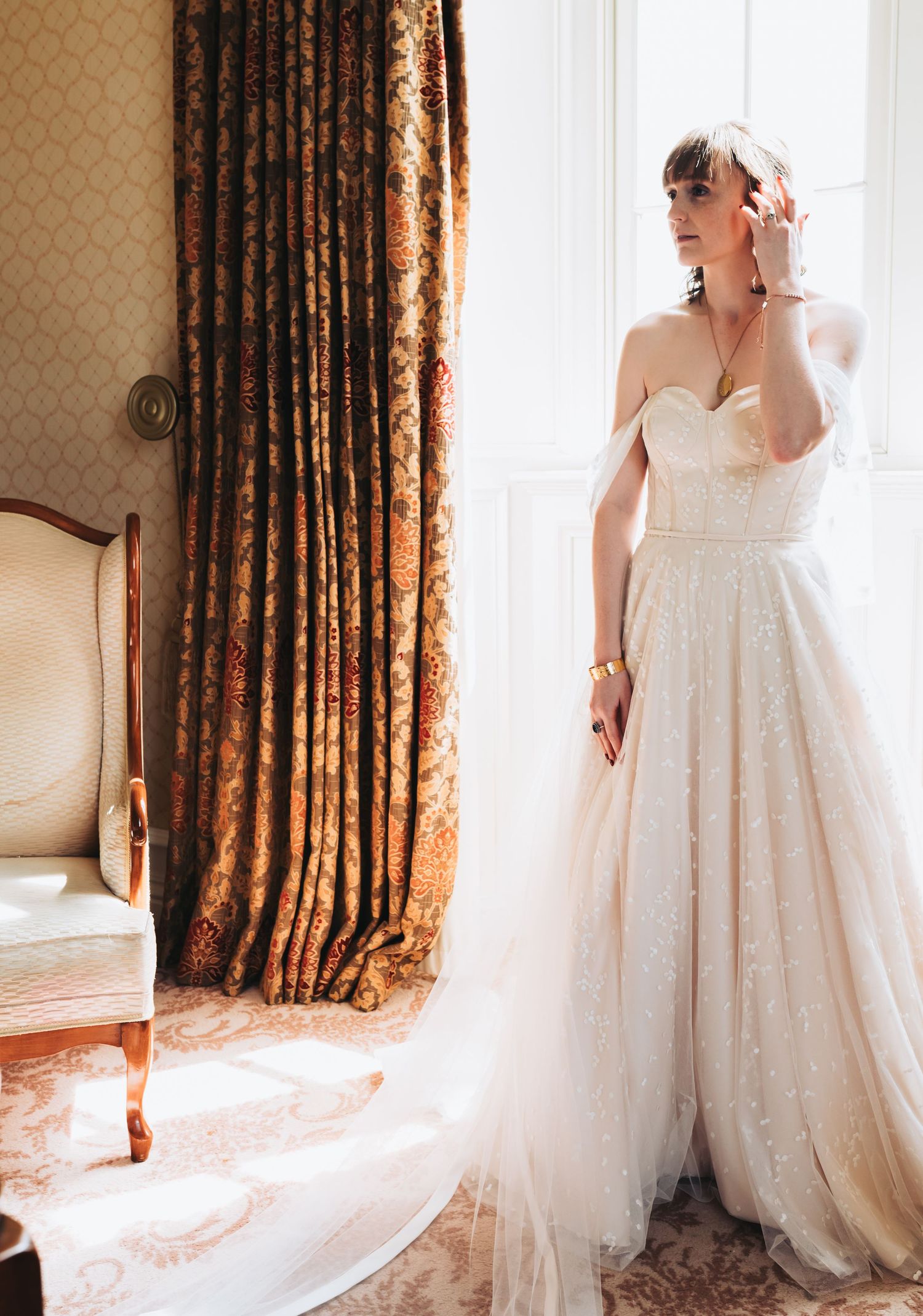Bride in strapless lace ball gown stands by window in ornate room with rich curtains.
