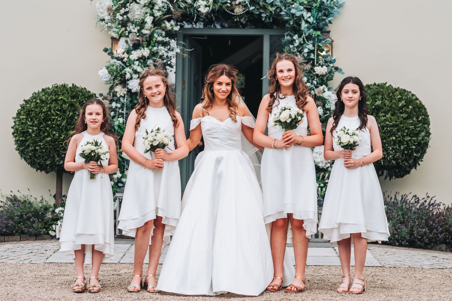 A bride poses with four young bridesmaids in white dresses holding bouquets outside a floral venue.