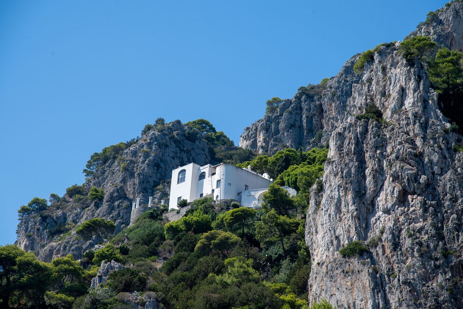 A white Mediterranean villa perched on a dramatic rocky cliff surrounded by lush green trees under blue sky.