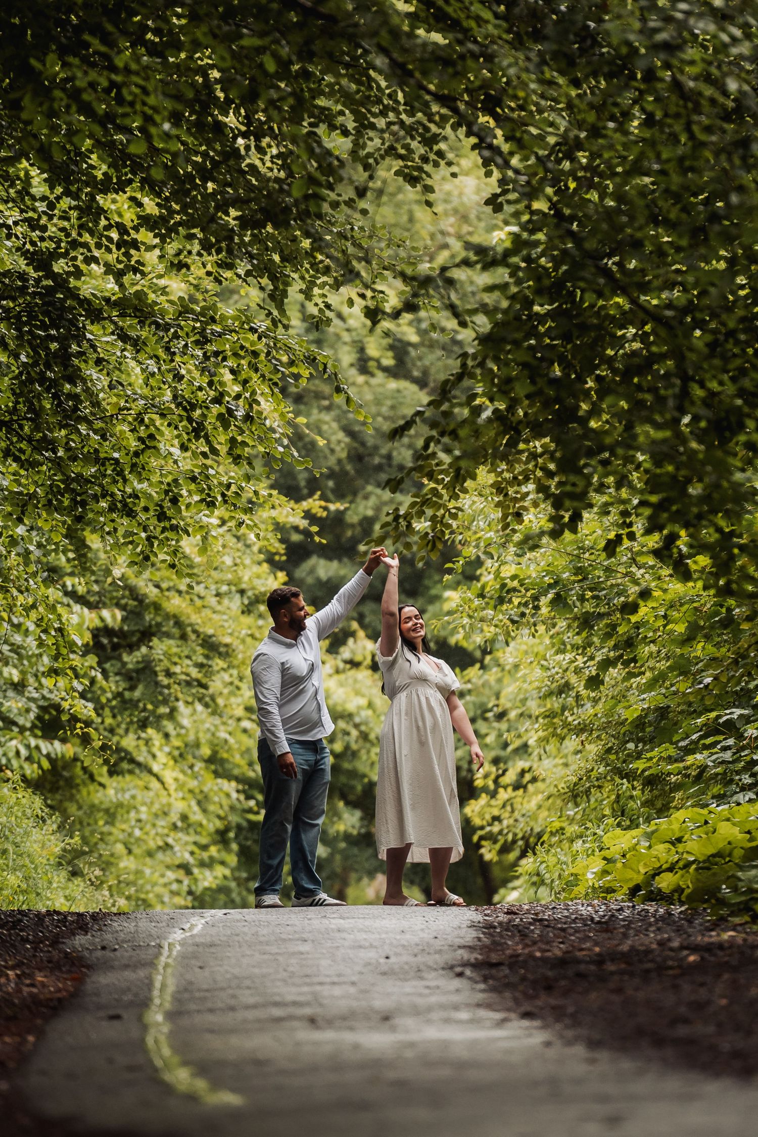 Couple dancing joyfully on a tree-lined path surrounded by lush green foliage.