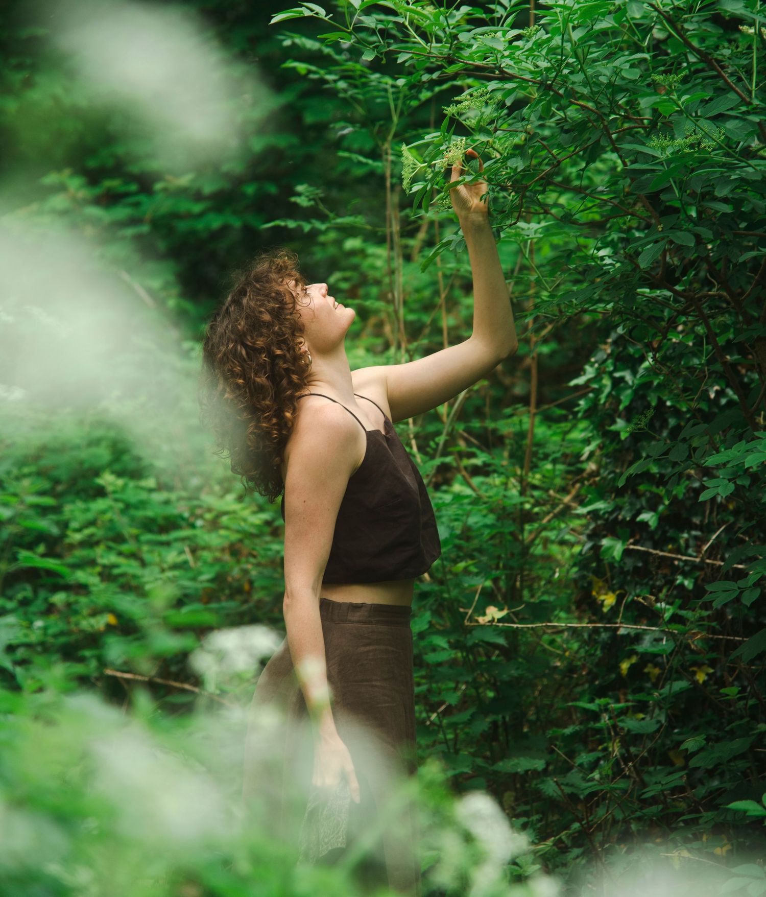 Curly-haired woman in black tank top reaching up to touch green leaves in forest.