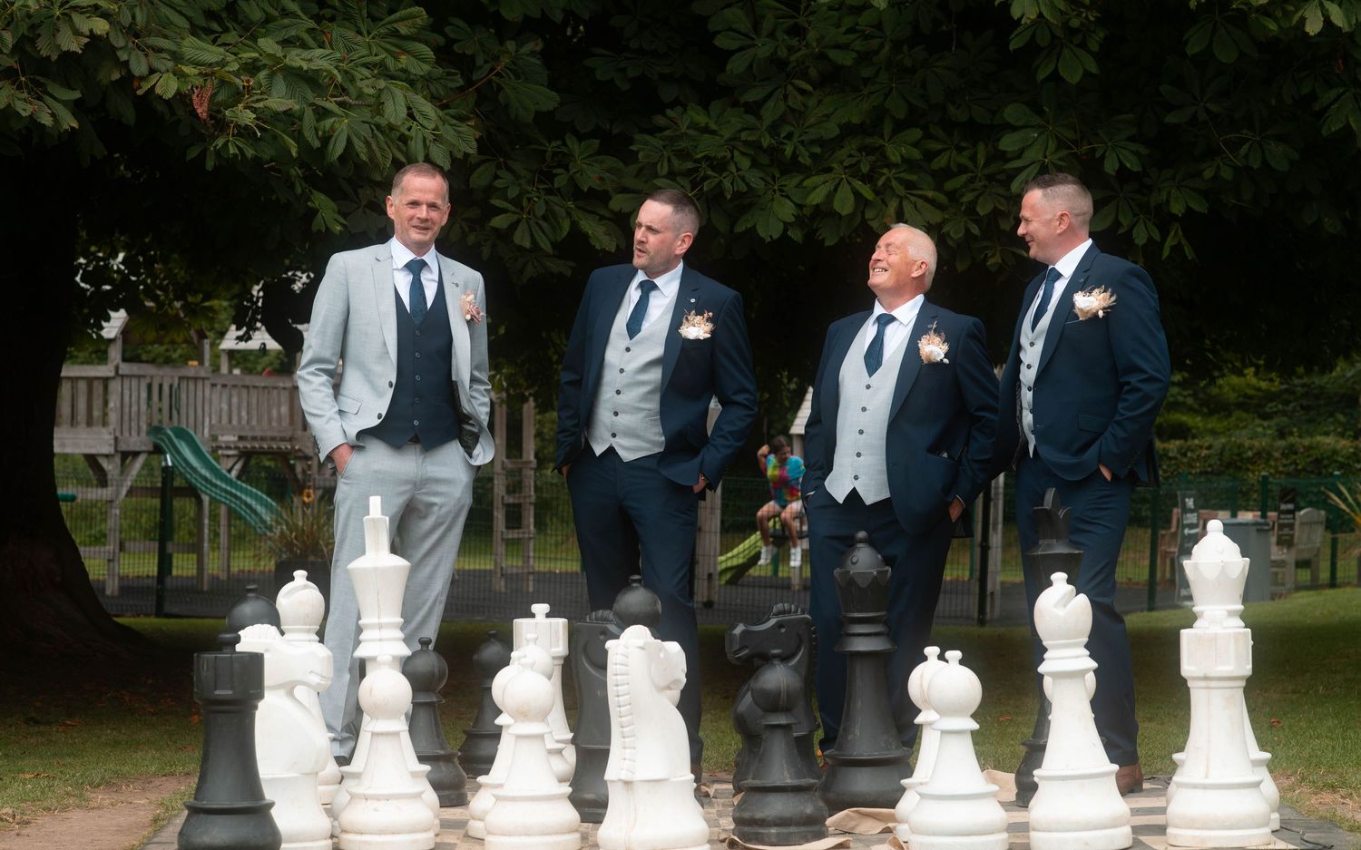 Four groomsmen in suits posing with giant outdoor chess pieces in garden.