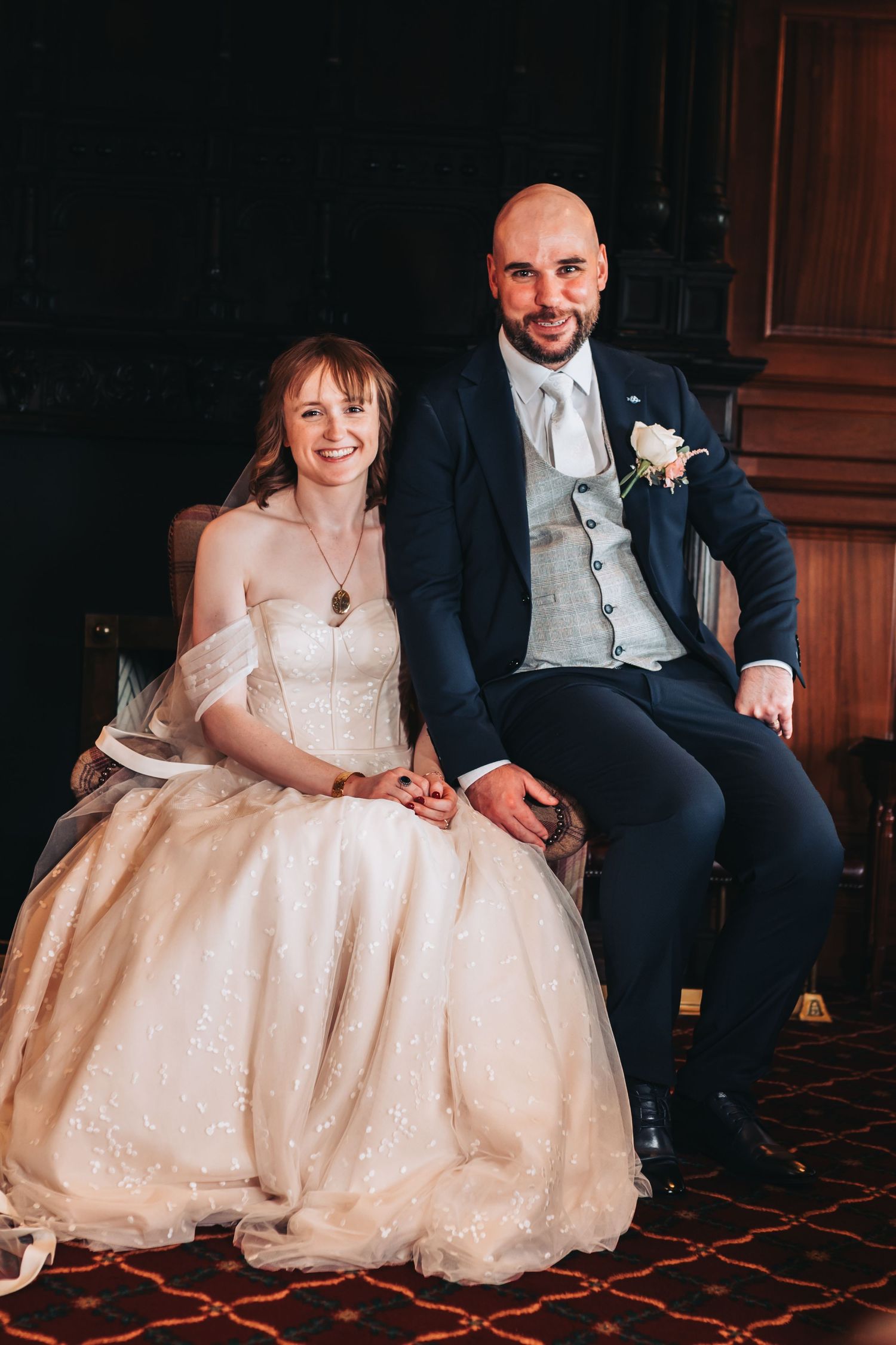 Smiling bride and groom sitting together, she in lace gown, he in navy suit with white boutonniere.
