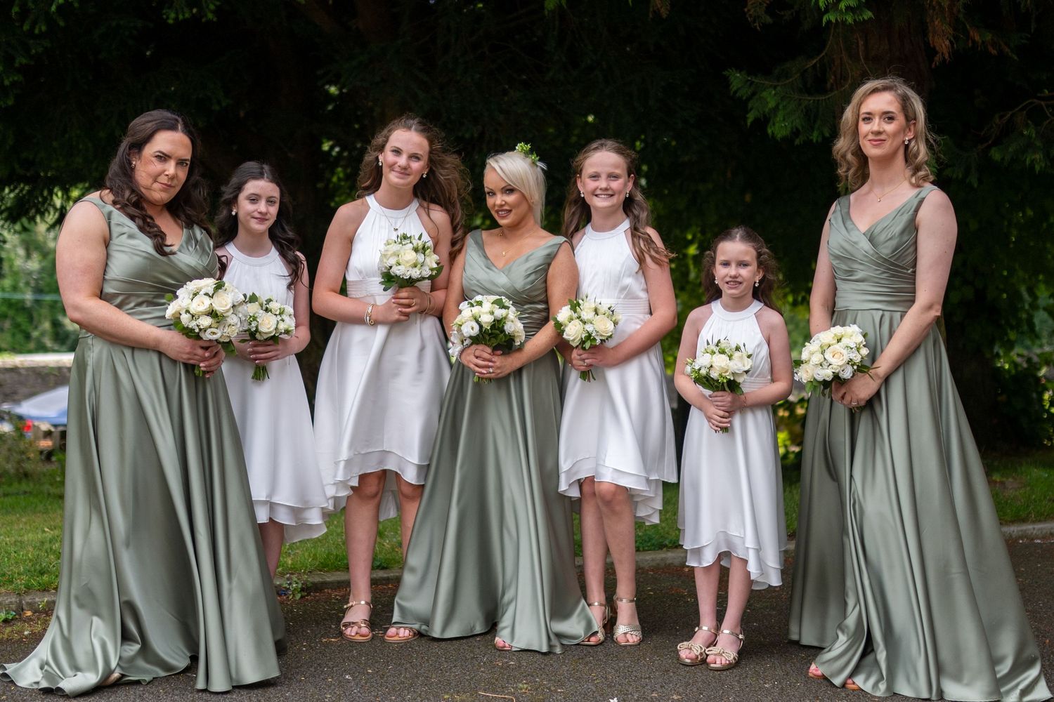 Bridesmaids in sage green gowns and flower girls in white dresses pose outdoors holding white bouquets at a wedding.