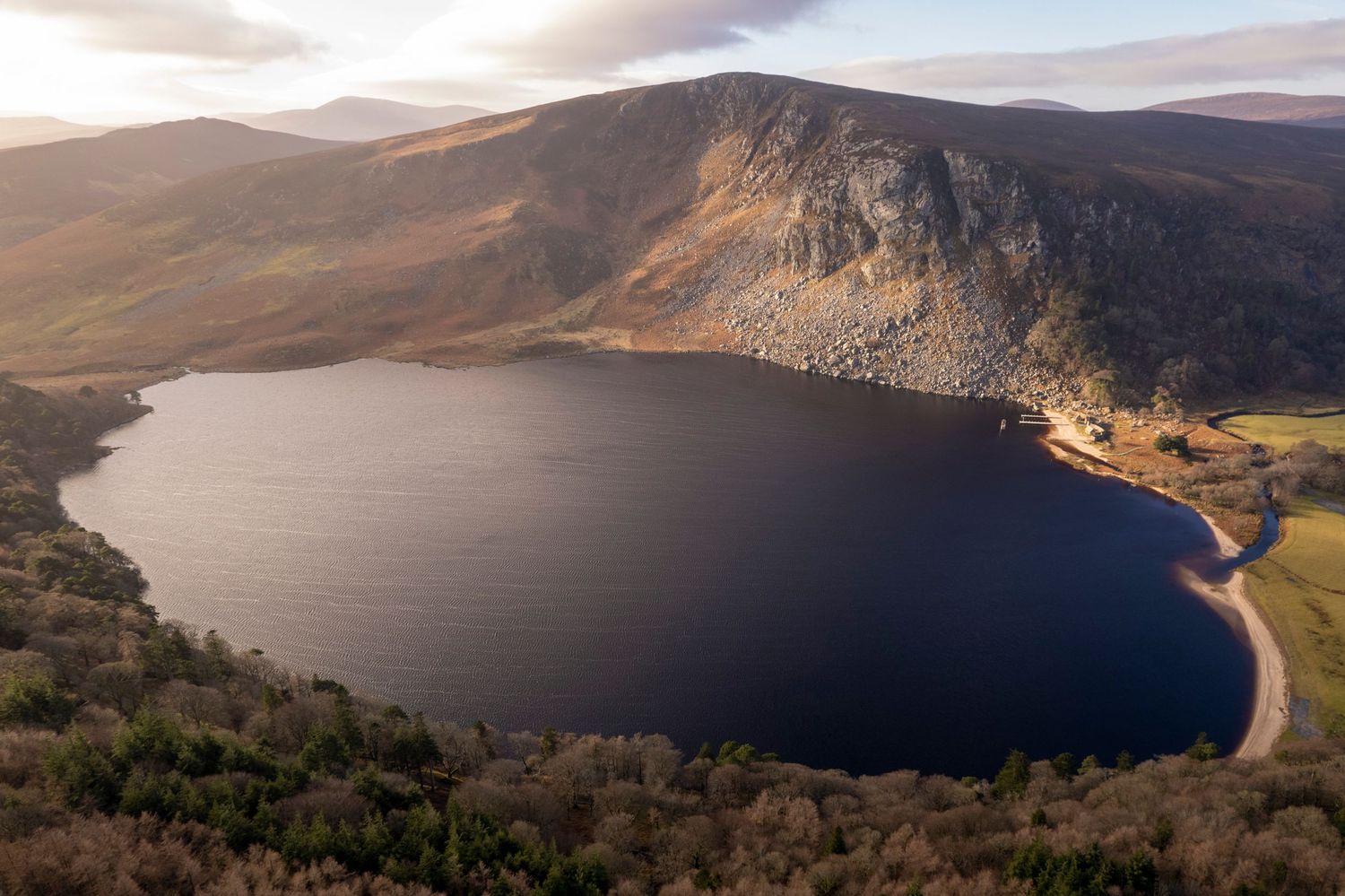 Aerial view of a dark glacial lake surrounded by mountains and autumn trees in Ireland.