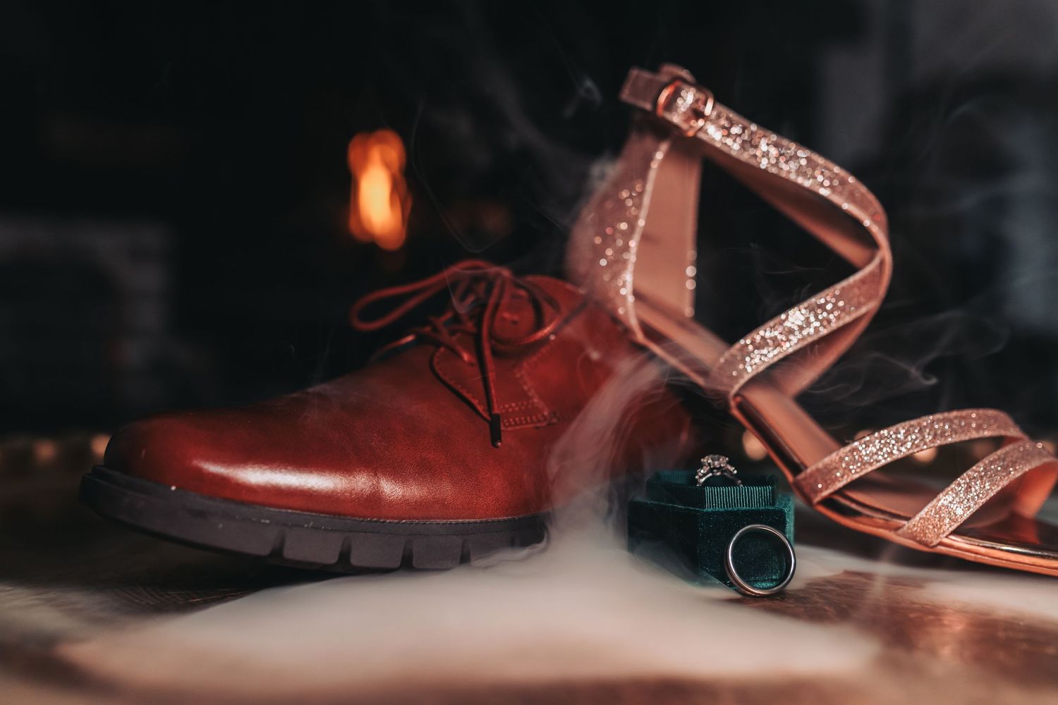 A polished brown leather shoe and a glittery heeled sandal with smoke and a perfume bottle nearby.