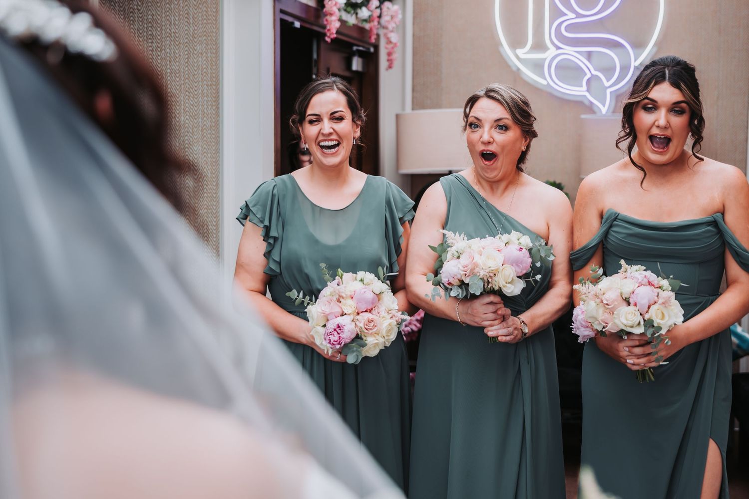 Three bridesmaids in green dresses react with joy and surprise seeing the bride, holding pink and white bouquets.