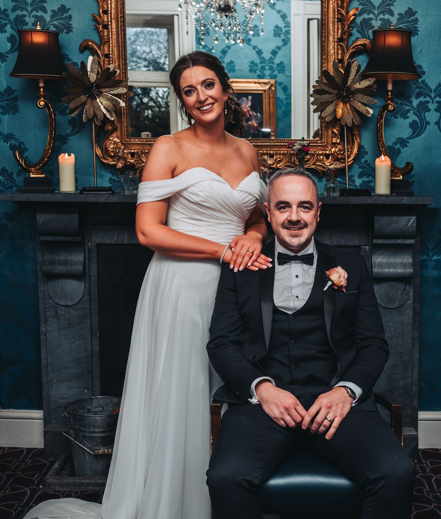 Elegant wedding couple posing in front of ornate gold fireplace, bride in off-shoulder gown, groom in black tuxedo.