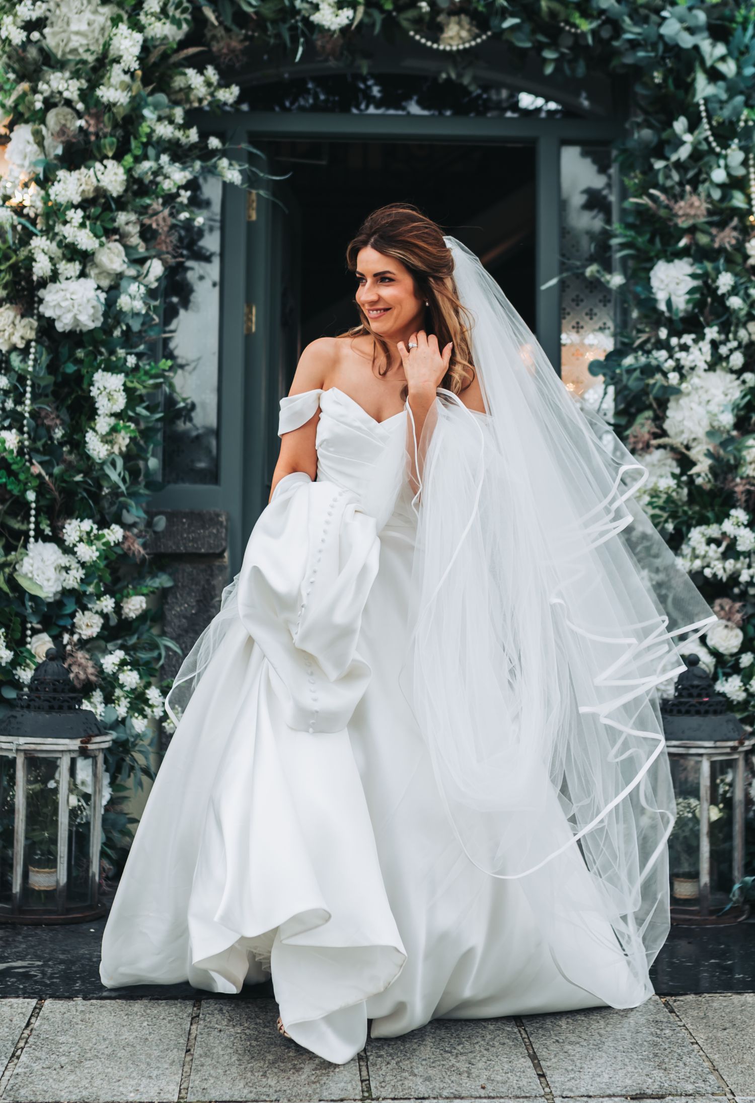 Bride in off-shoulder white gown with veil poses before floral arch of white blooms and greenery.
