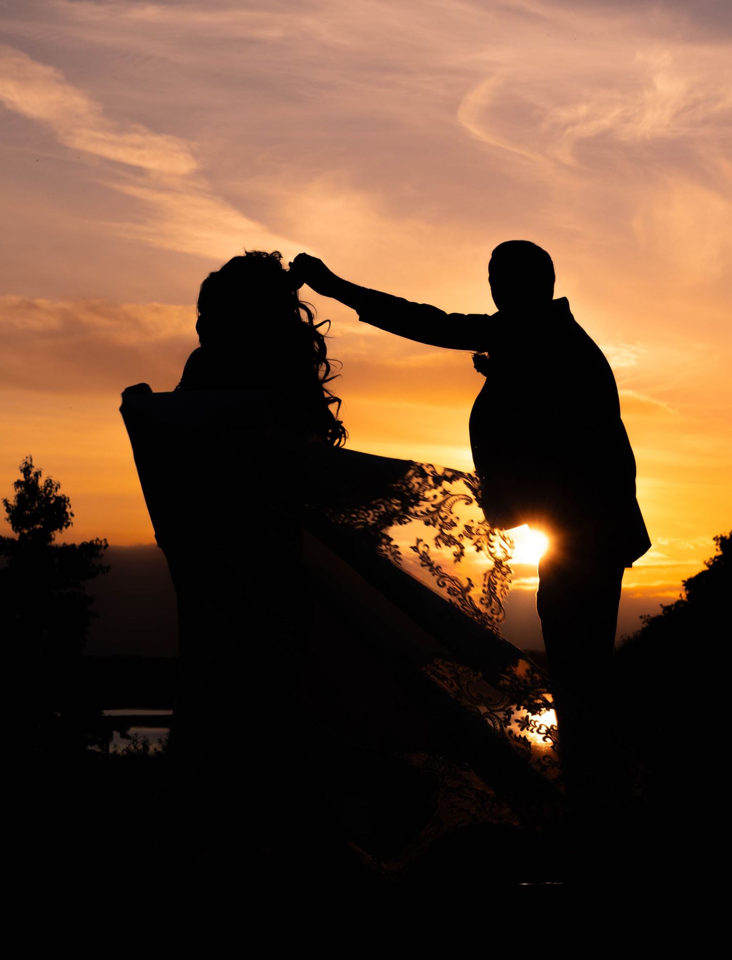 Silhouettes of a couple against a dramatic sunset sky with the veil flowing in the wind.