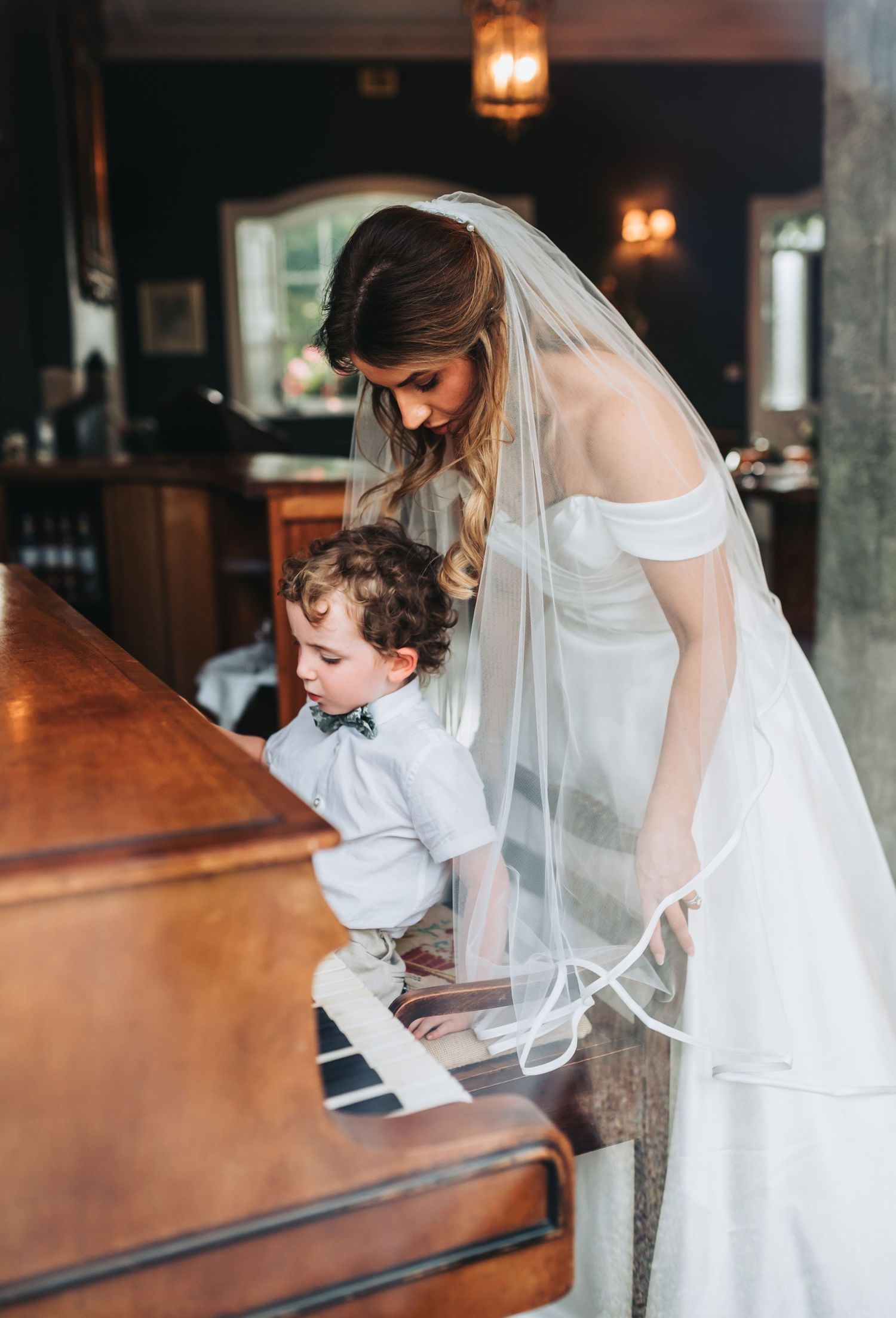 Bride in off-shoulder gown and veil stands with young boy playing piano at wedding venue.