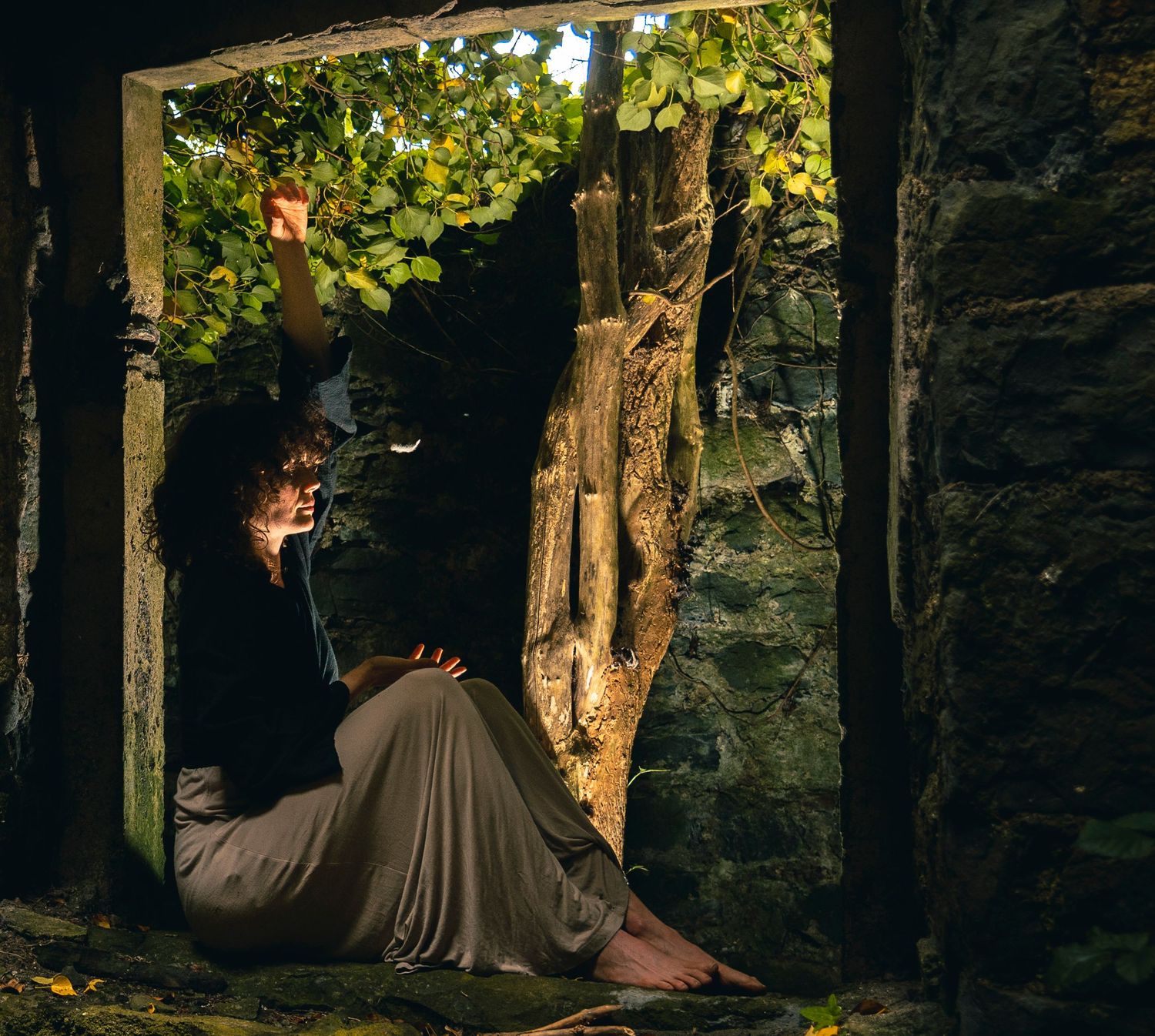 Woman in flowing skirt sitting in sunlit stone archway reading among green vines and trees.