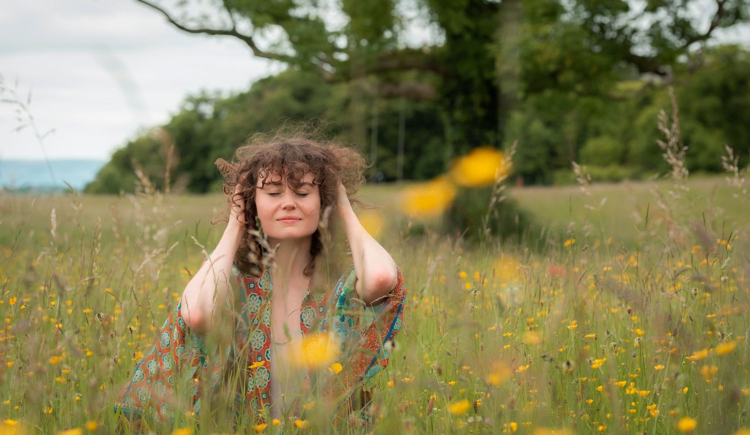 Woman with curly hair standing in yellow wildflower field with eyes closed and hands on head.