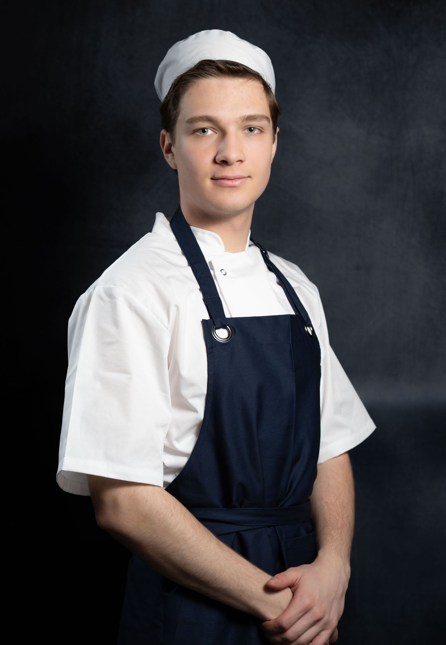 Young chef in white jacket and navy blue apron with chef hat poses professionally against dark backdrop.