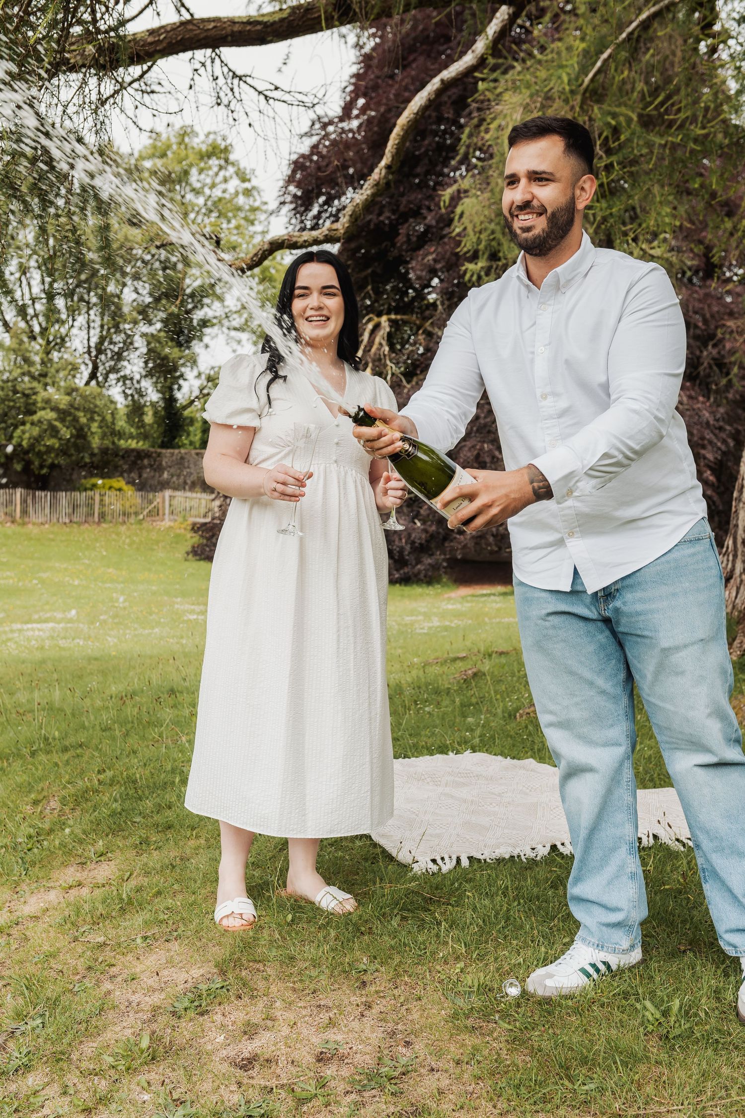 Happy couple celebrating outdoors, man popping champagne bottle while woman in white dress holds a glass in a garden.