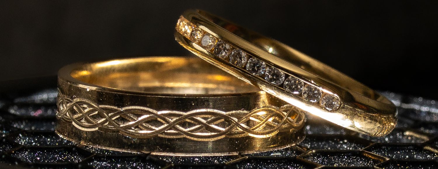 Two gold wedding bands with intricate Celtic designs and diamond accents on dark textured surface.