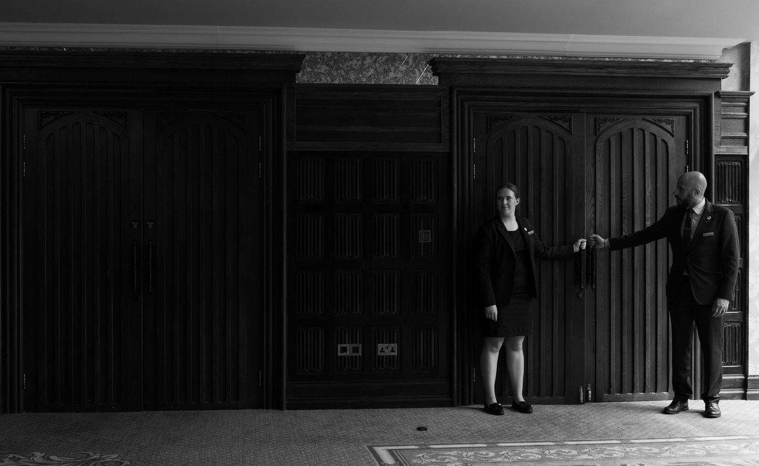 Black and white photo of two people standing by large wooden doors in formal attire.