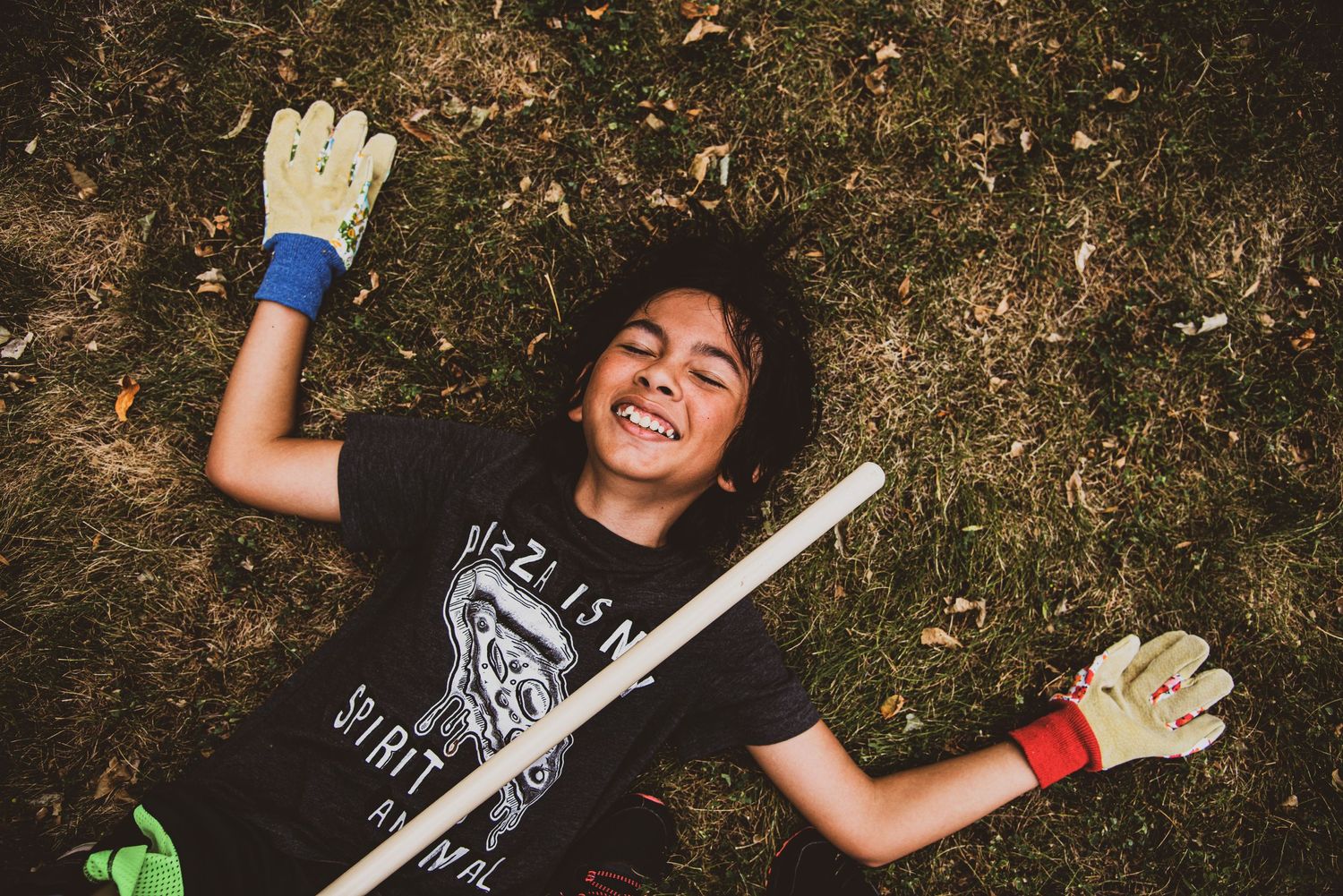Joyful child in gardening gloves laughing while lying on green grass with a stick