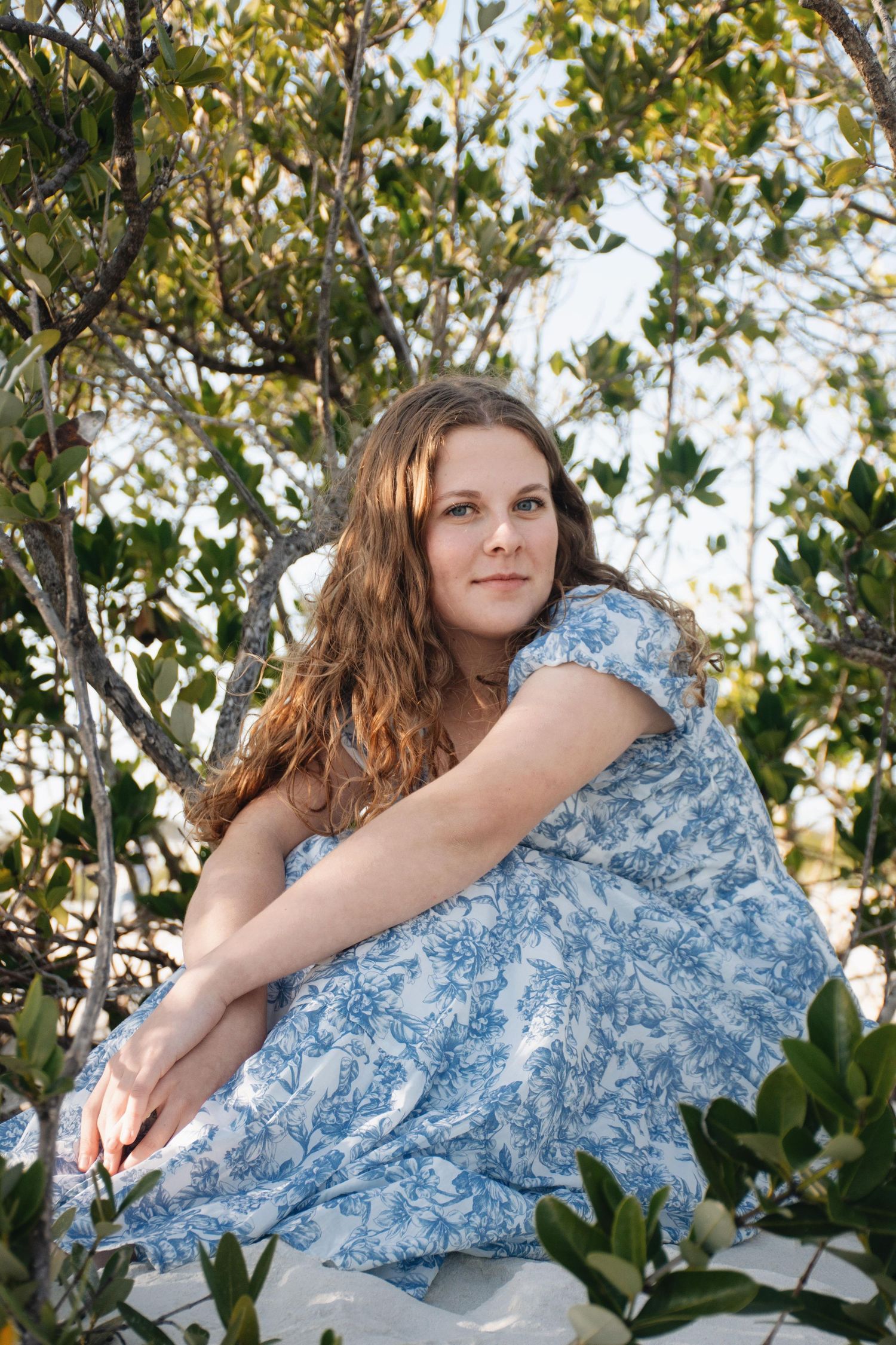 A person in a blue floral dress reclines against a tree branch surrounded by green foliage.
