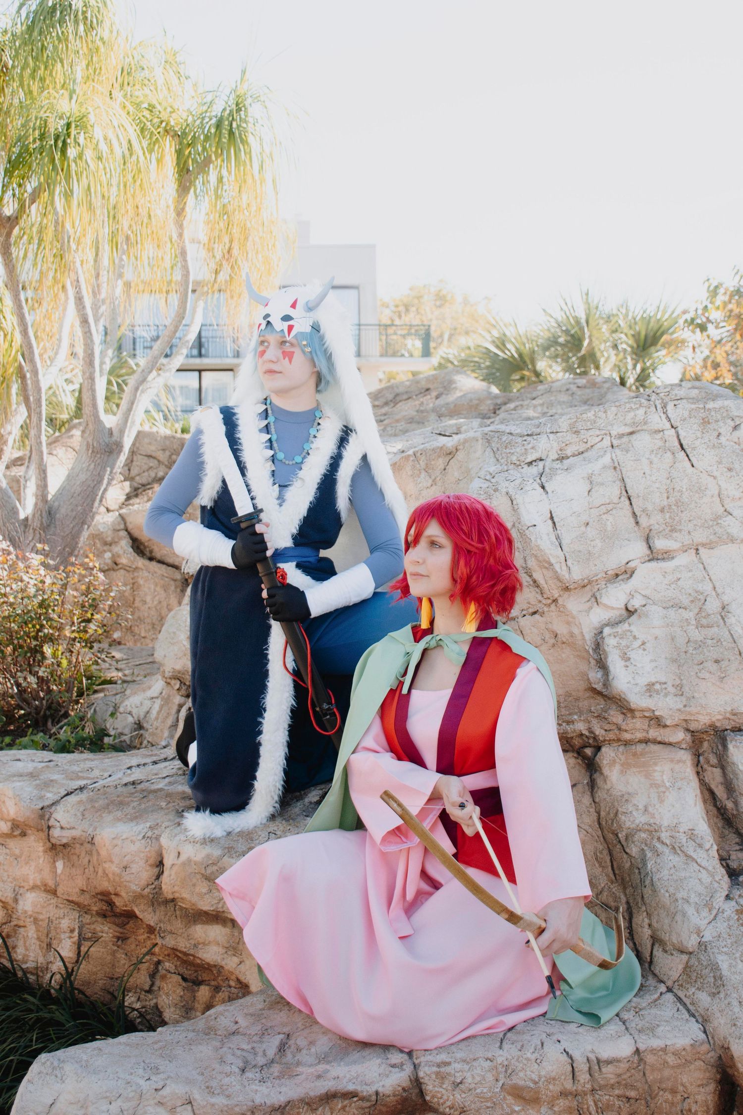 A cosplay scene with two characters in pink and blue outfits posing together against a rocky outdoor backdrop with palm trees.