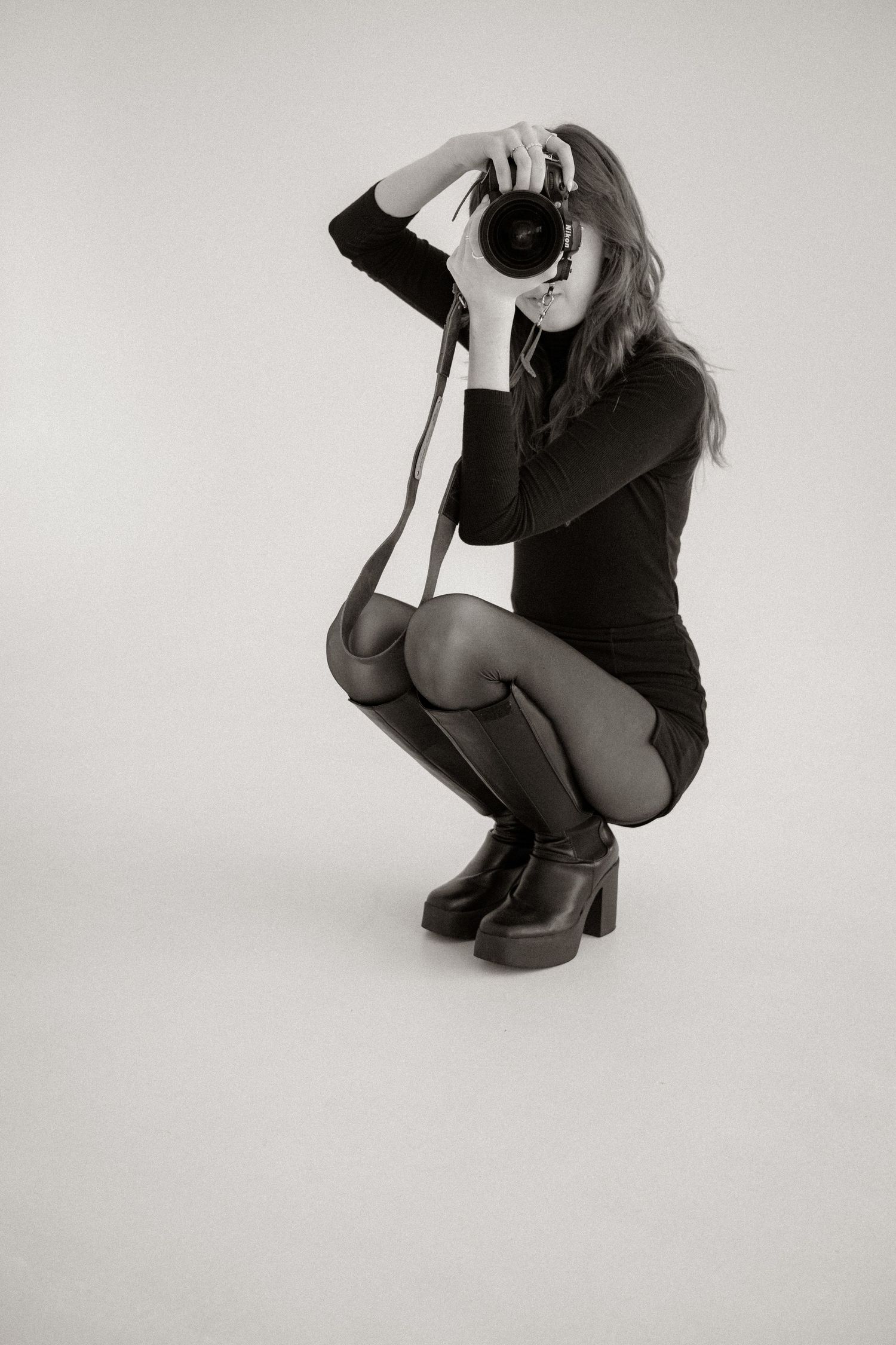 Black and white photo of a woman crouching in a black dress and boots, holding a DSLR camera up to her eye.