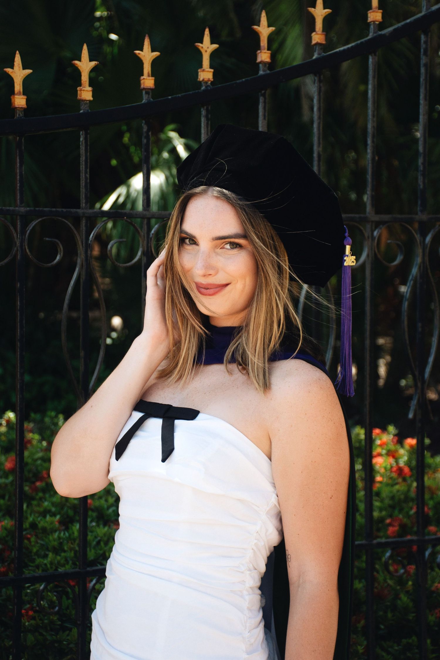 Graduate in white dress and cap poses by decorative iron fence during graduation photos.