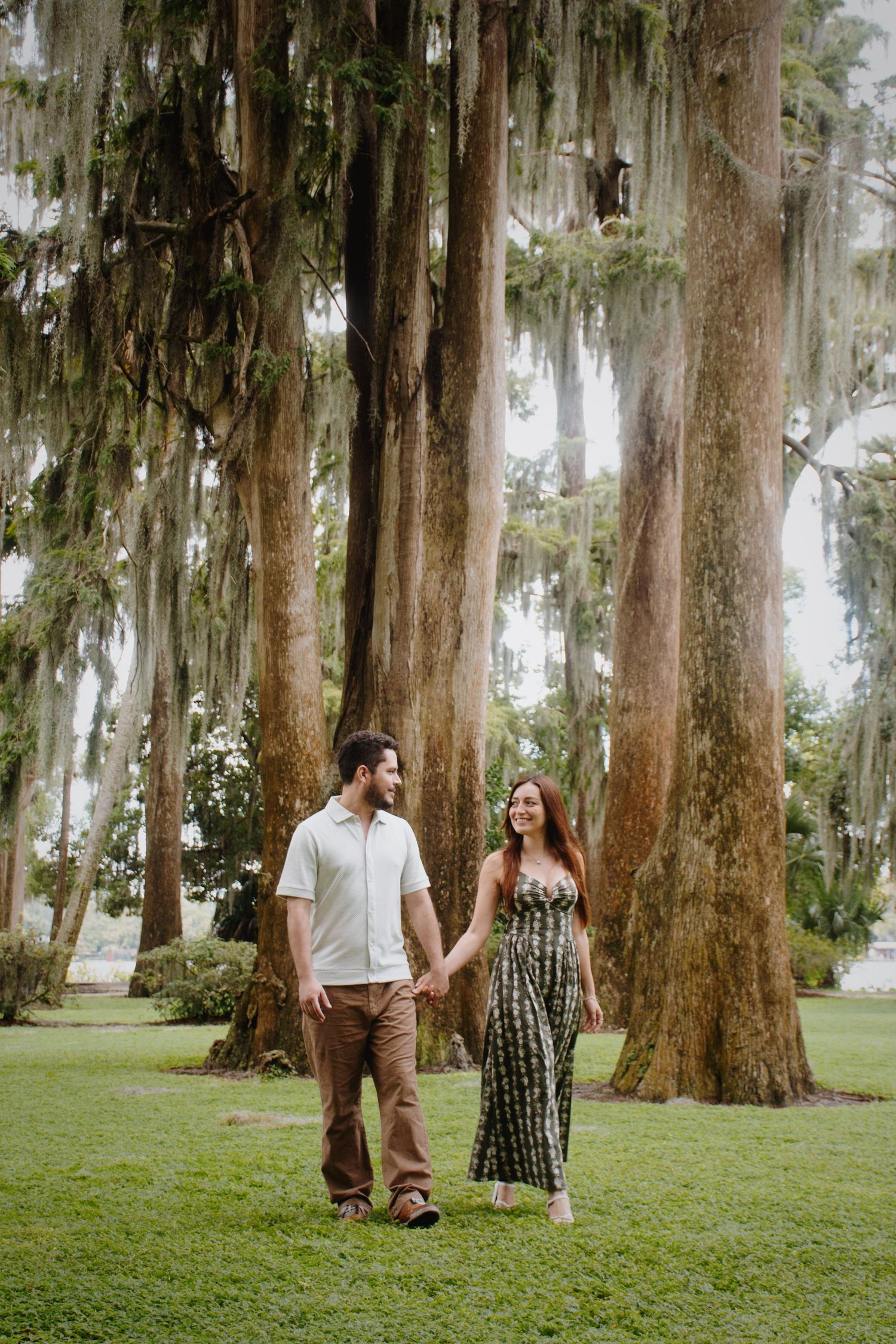 Couple walking hand in hand through a scenic park with Spanish moss draped trees.